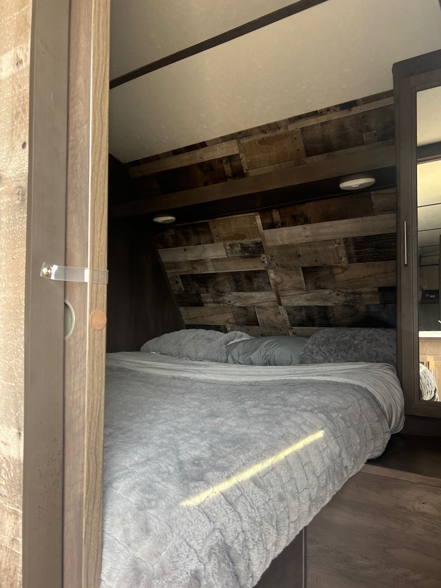 Cozy RV rental bedroom with a rustic wood headboard, plush gray bedding, and mirrored closet. Ready for your trip!. Aspen Trail Aspen Trail Trailer 2021