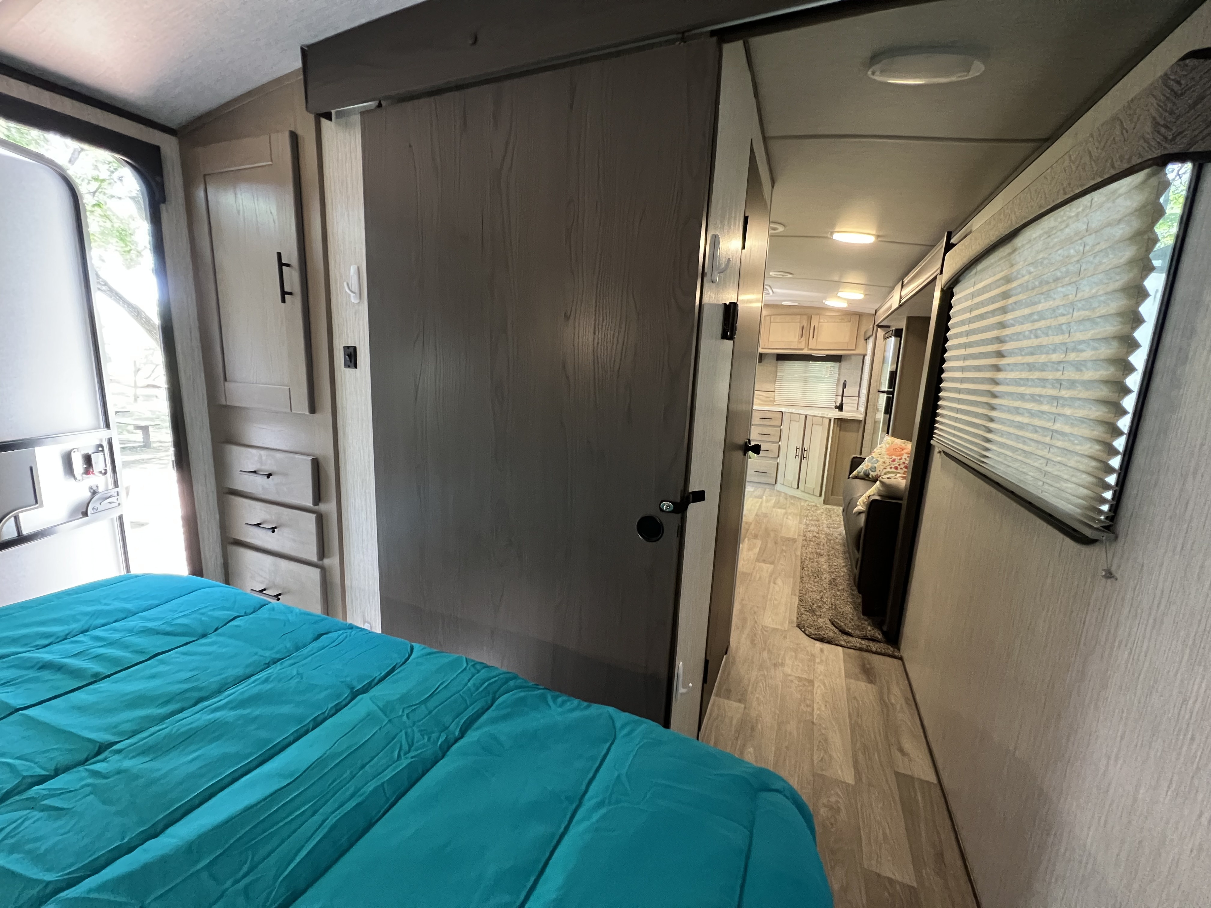 RV rental interior: Teal bed in bedroom, open door to outdoors, leading to kitchen and living area with light wood finishes.. Sundance 242RK 2020