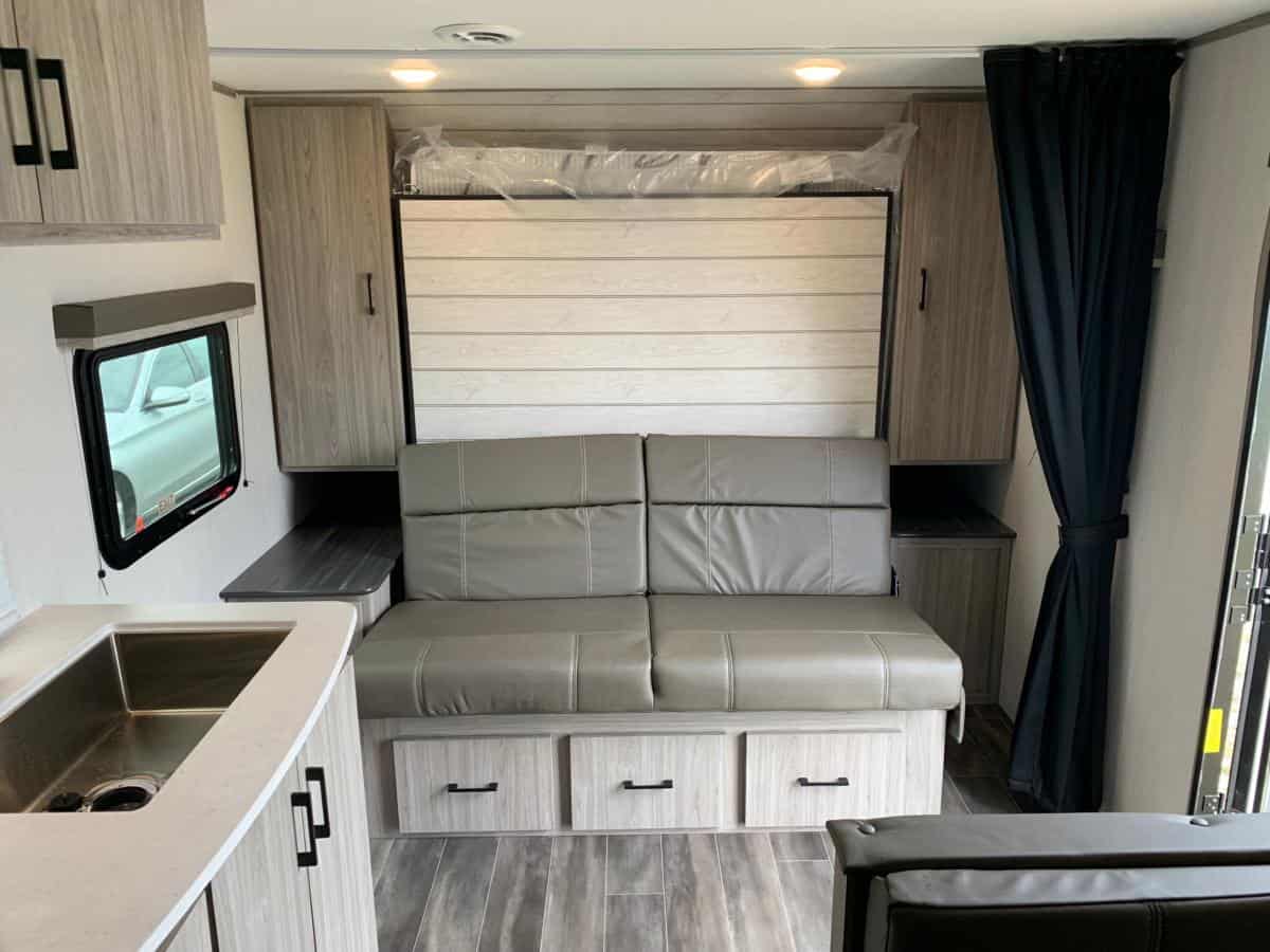 Cozy RV rental interior featuring a versatile light gray sofa, pull-down bed, compact kitchen, and wood-look storage.. Keystone RV Passport SL 2021