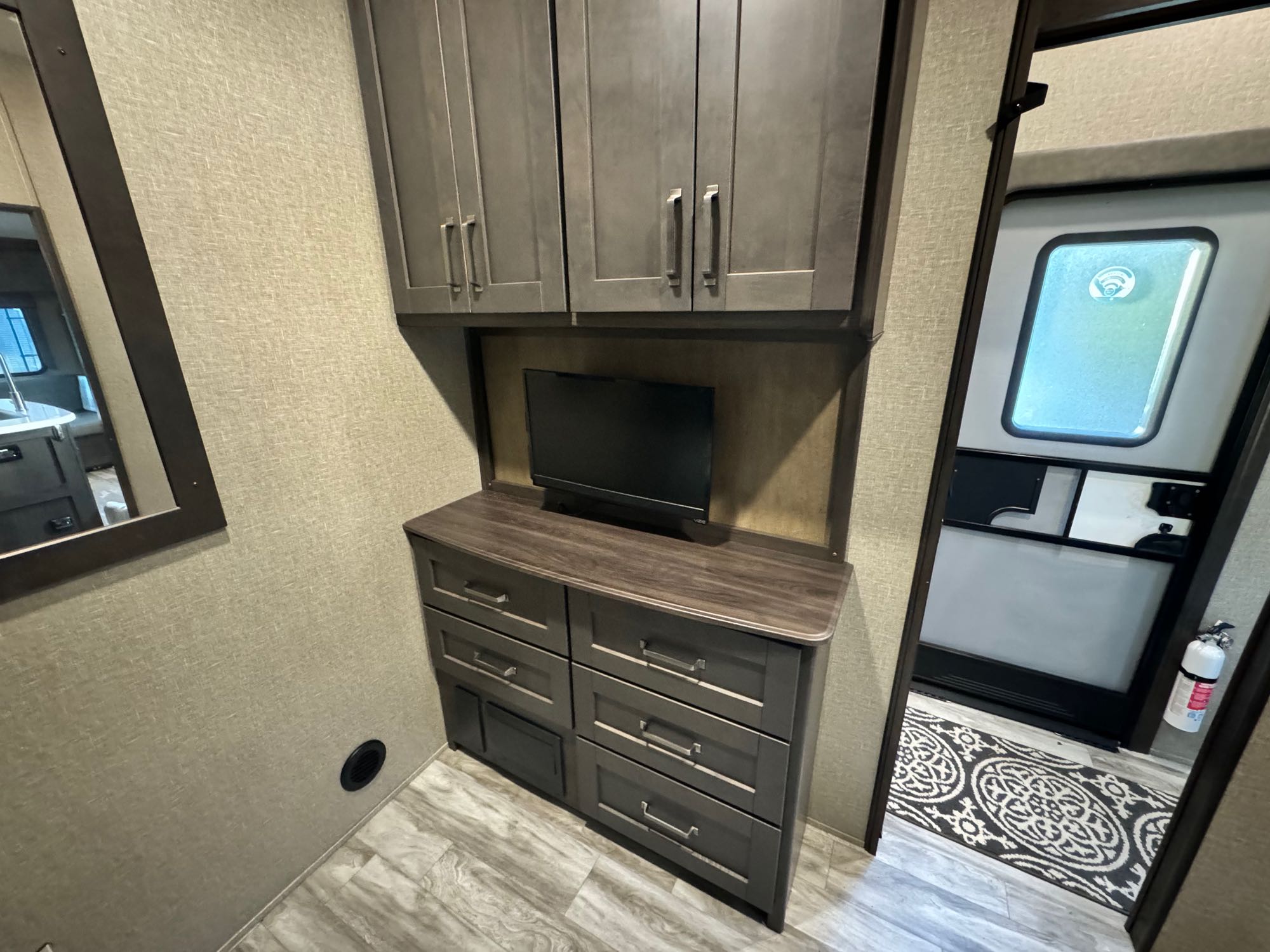 Inside this modern rv rental, you see a TV atop drawers, overhead cabinets, a mirror, & an exit door with a fire extinguisher.. Grand Design Reflection 2022