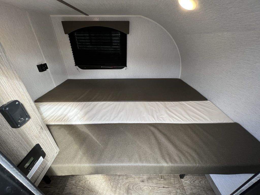 Explore a cozy RV rental interior: a modular bed with dark and white cushions, window, and controls for your trip.. Sunset Park RV SunRay 2023