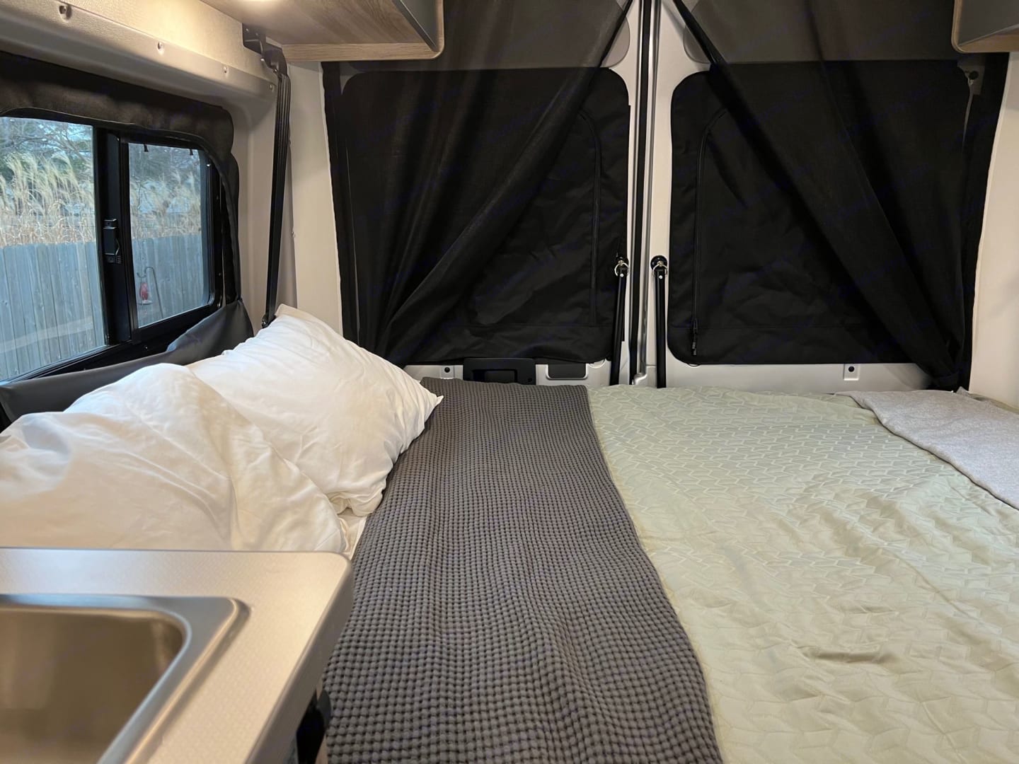 Cozy RV rental interior features a made bed, window with view, sink, and dark privacy shades on back windows.. Winnebago Solis 2023