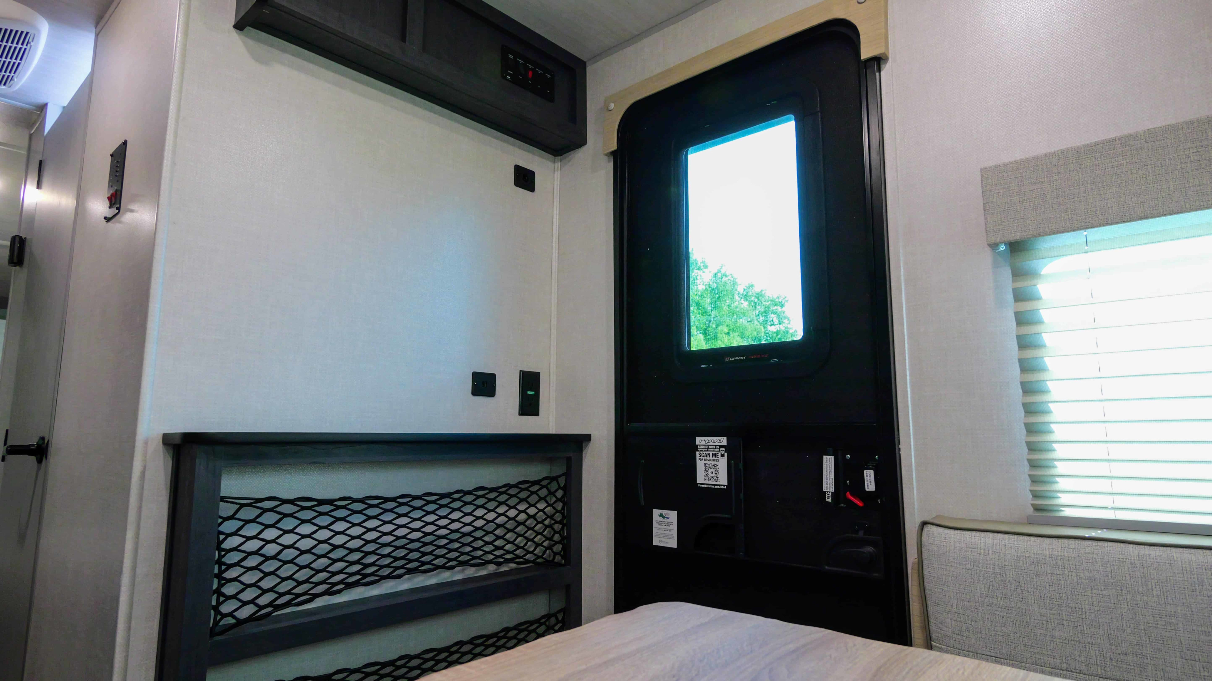 An RV rental's interior reveals a bed with mesh storage, a door with window view, and overhead controls.. Forest River R-Pod 190 2024