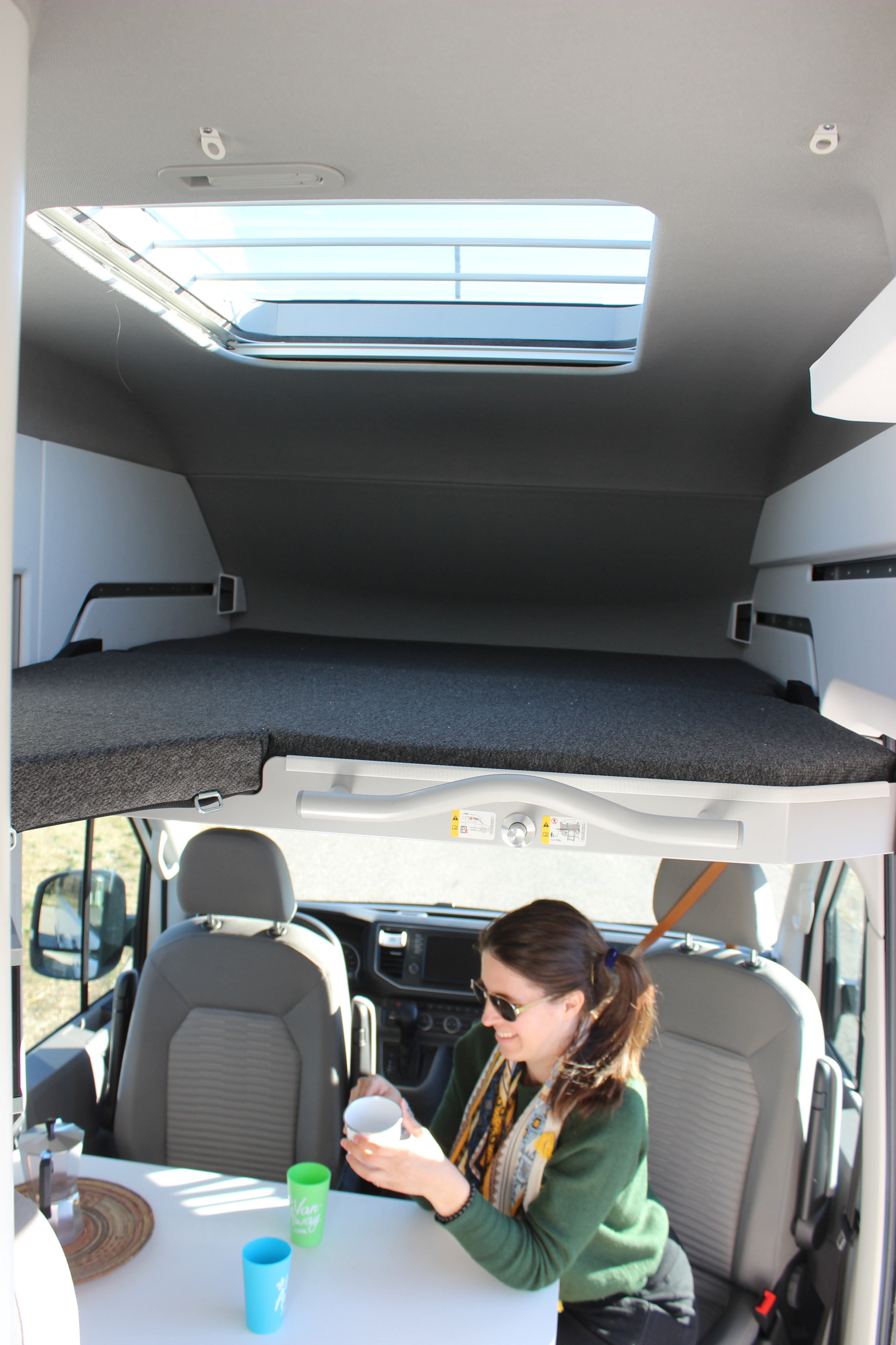 Cozy RV rental interior with an upper bed, skylight, and a woman enjoying coffee at the dining area.. Volkswagen Grand California 600 2023