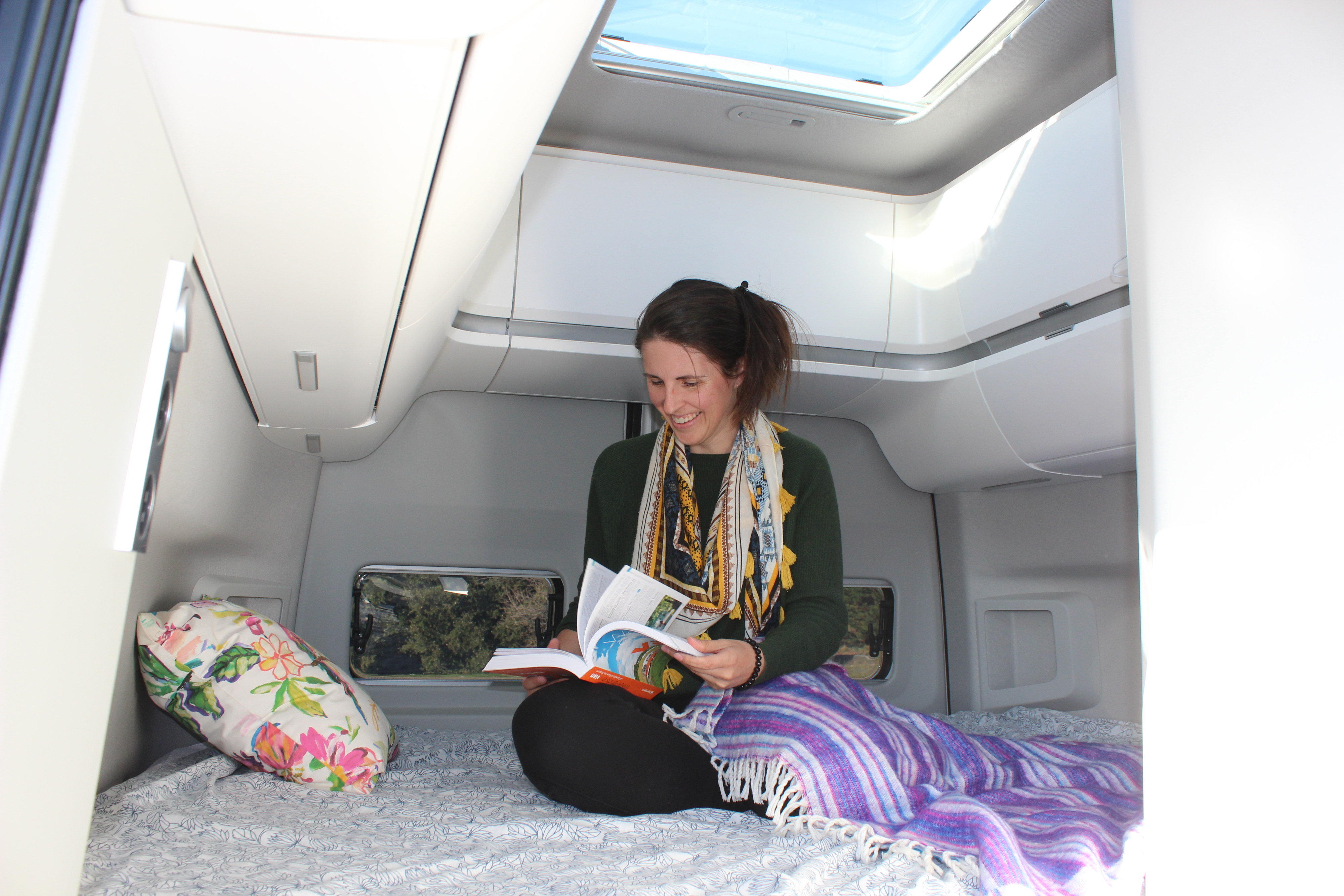 Cozy up in your RV rental! A woman reads in a bright, comfortable camper van interior with a skylight overhead.. Volkswagen Grand California 600 2023