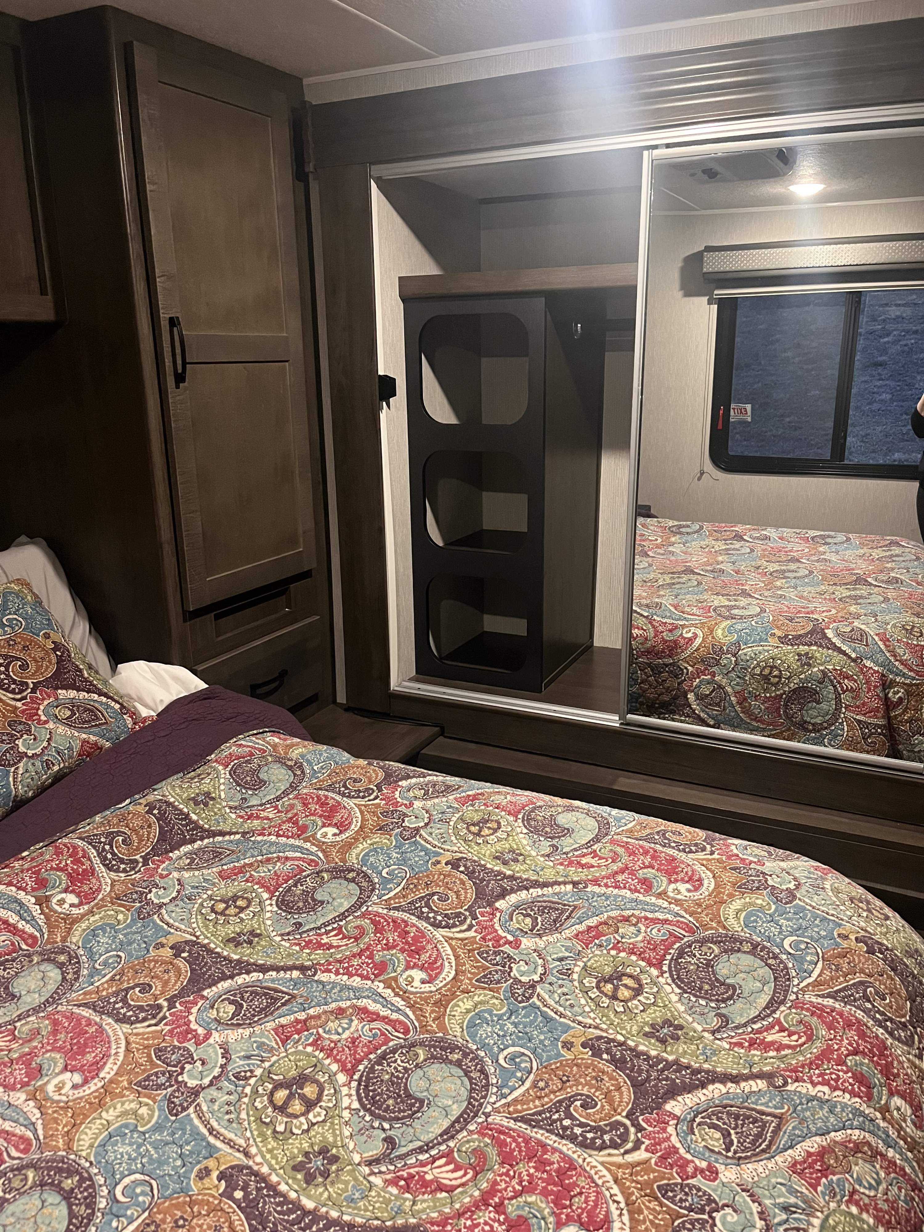 Cozy RV rental bedroom with a colorful paisley-patterned bedspread, dark wood cabinets, and a large mirror reflecting the bed and a window.. Coachmen Leprechaun Premier 2023
