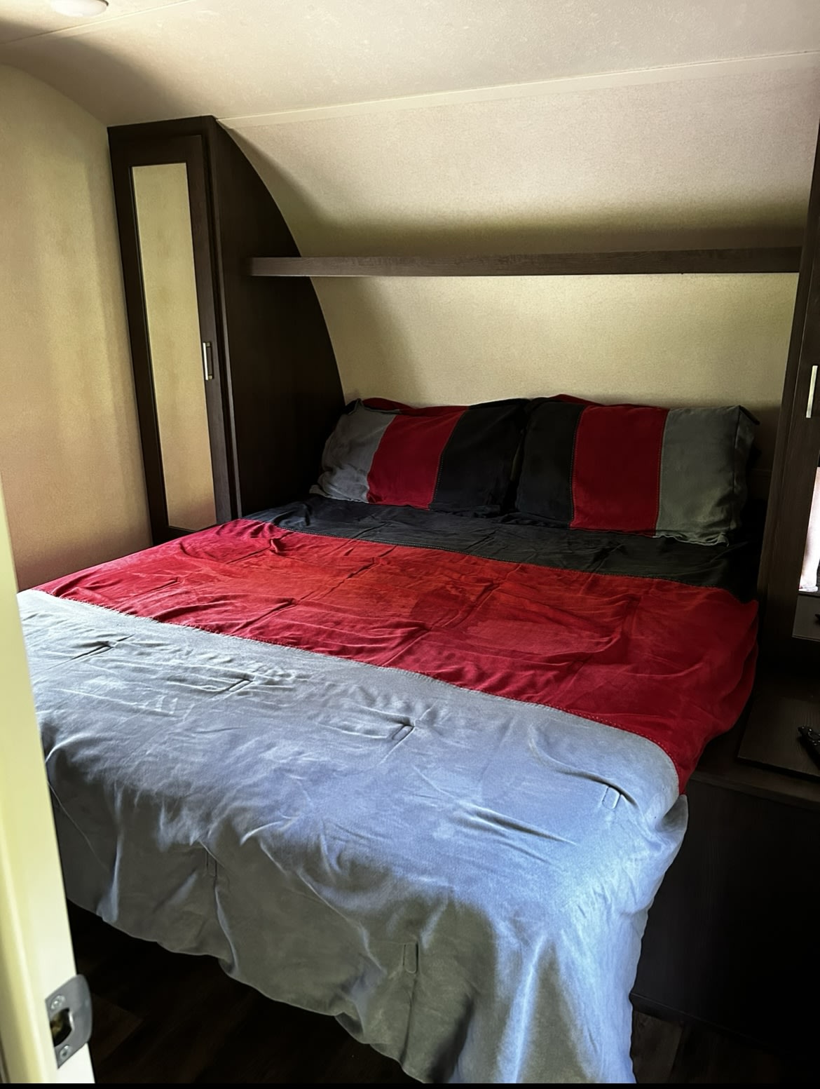 Inside an RV rental: a compact bedroom featuring a bed with red & grey bedding, built-in storage, and a shelf.. Forest River Wildwood 2019