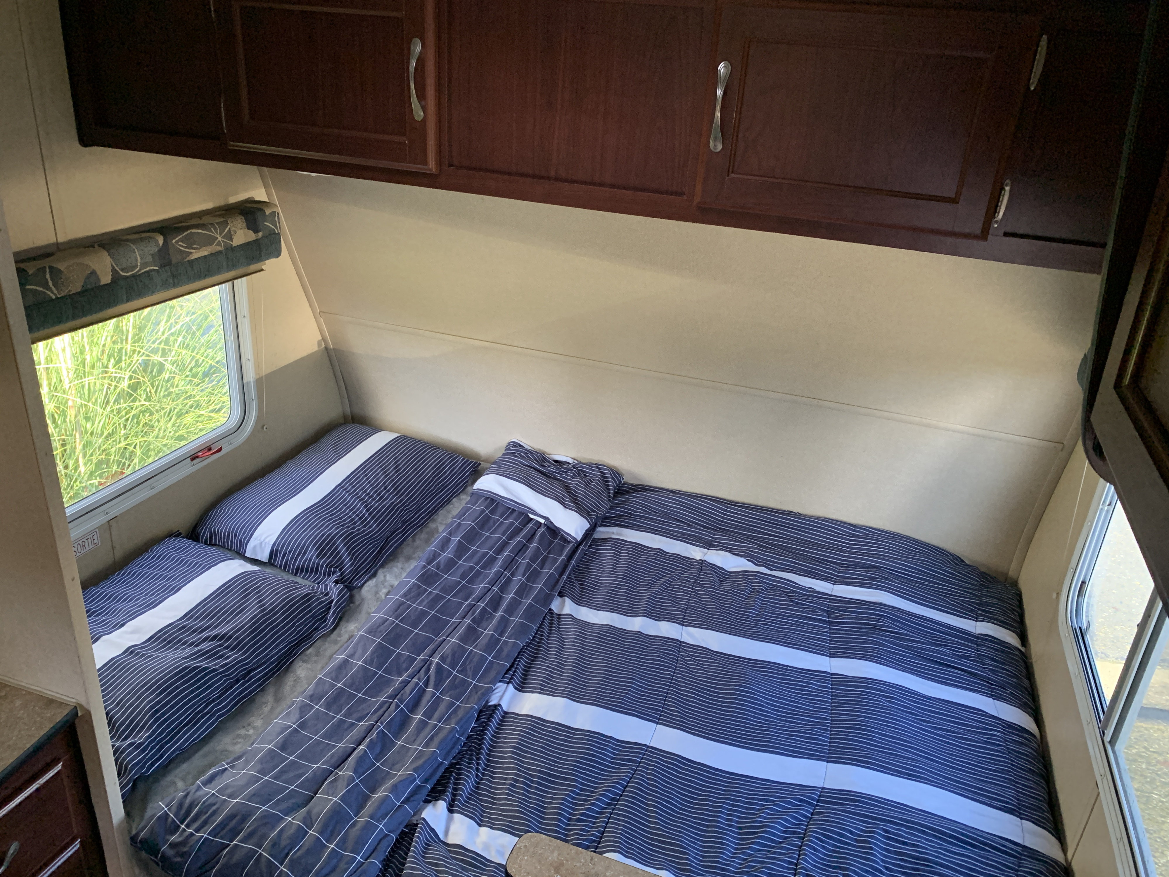 Interior of an RV rental bedroom featuring a bed with blue striped bedding, pillows, overhead cabinets, and a window displaying green foliage.. Takena Takena Trailer 2010