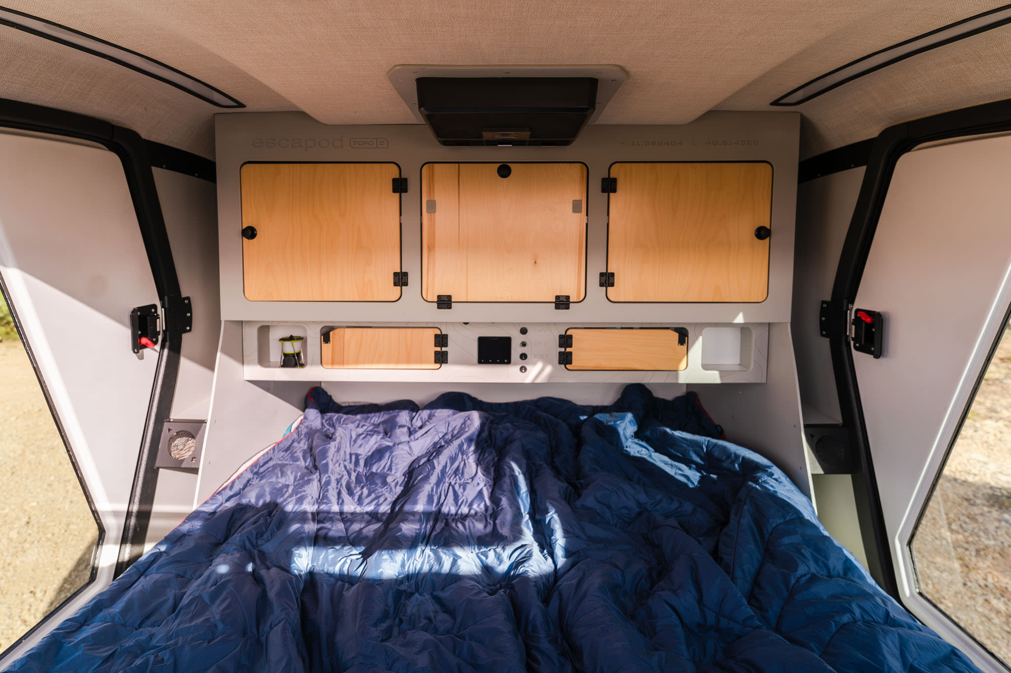 Compact RV rental interior: A comfortable sleeping area with a blue comforter, overhead wooden cabinets, and integrated controls for adventures.. Escapod TOPO2 Voyager 2024