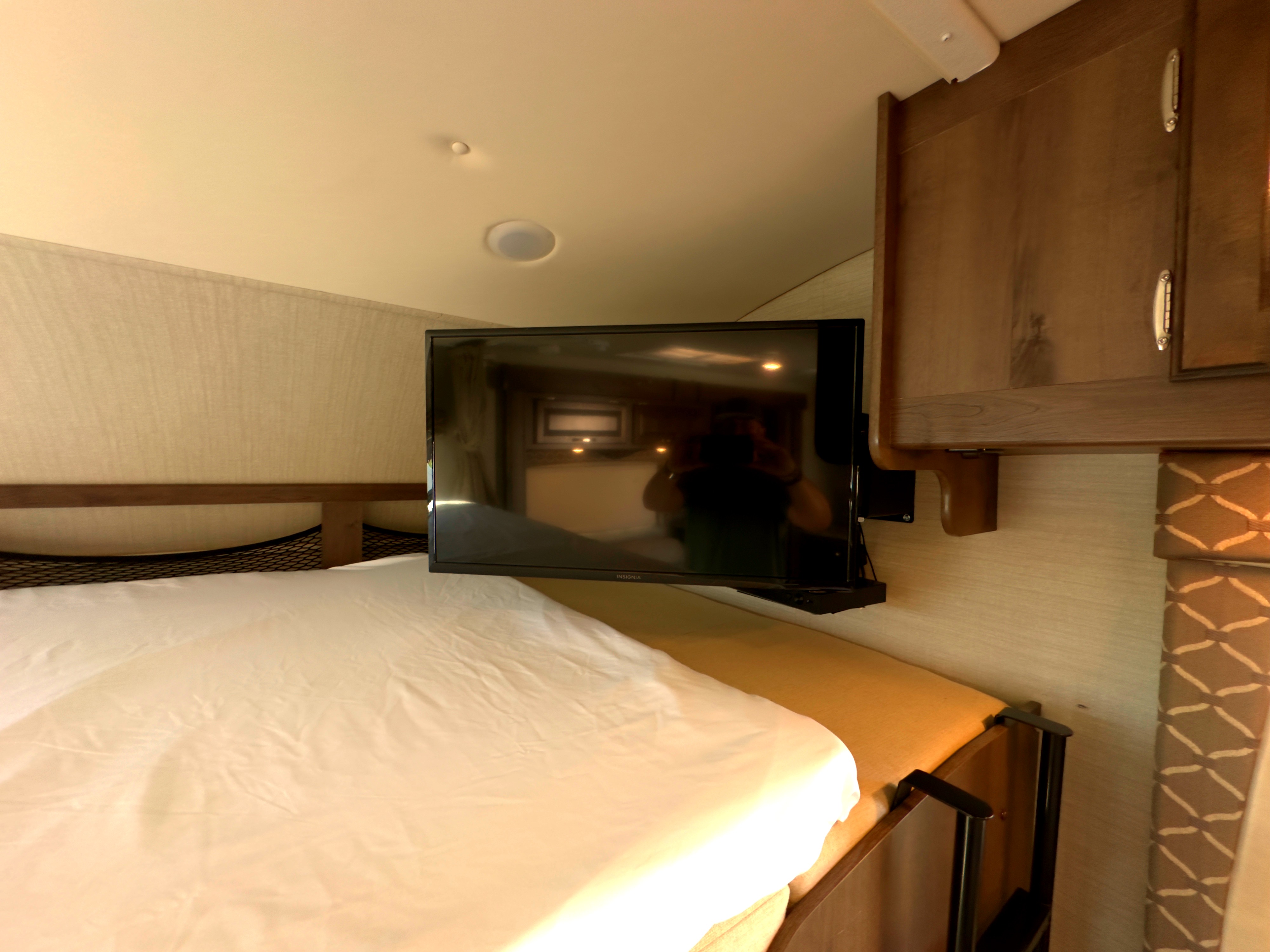 Interior of an RV rental: a bed with white sheets, wall-mounted TV, and wooden overhead cabinets for storage.. Jayco Greyhawk 2018