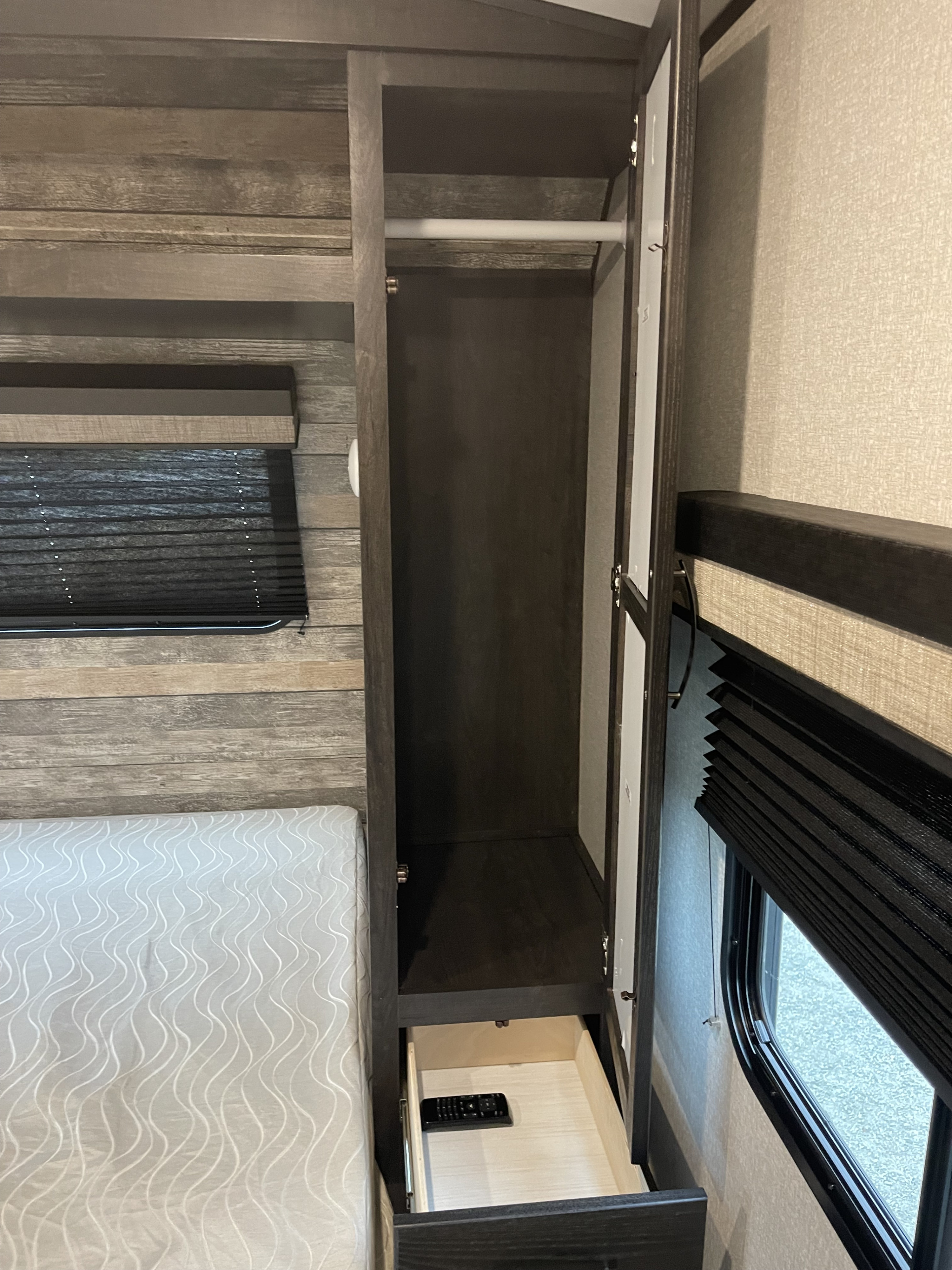 Inside an RV rental: a bedroom with an open wardrobe, hanging rod, drawer holding a remote, bed, and window with blind.. Forest River Surveyor Legend 2021