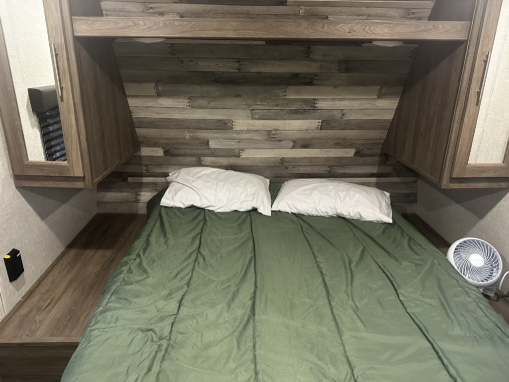 This RV rental features a cozy, compact bedroom with a bed, overhead cabinets, and wood paneling.. Crossroads RV Zinger 2023