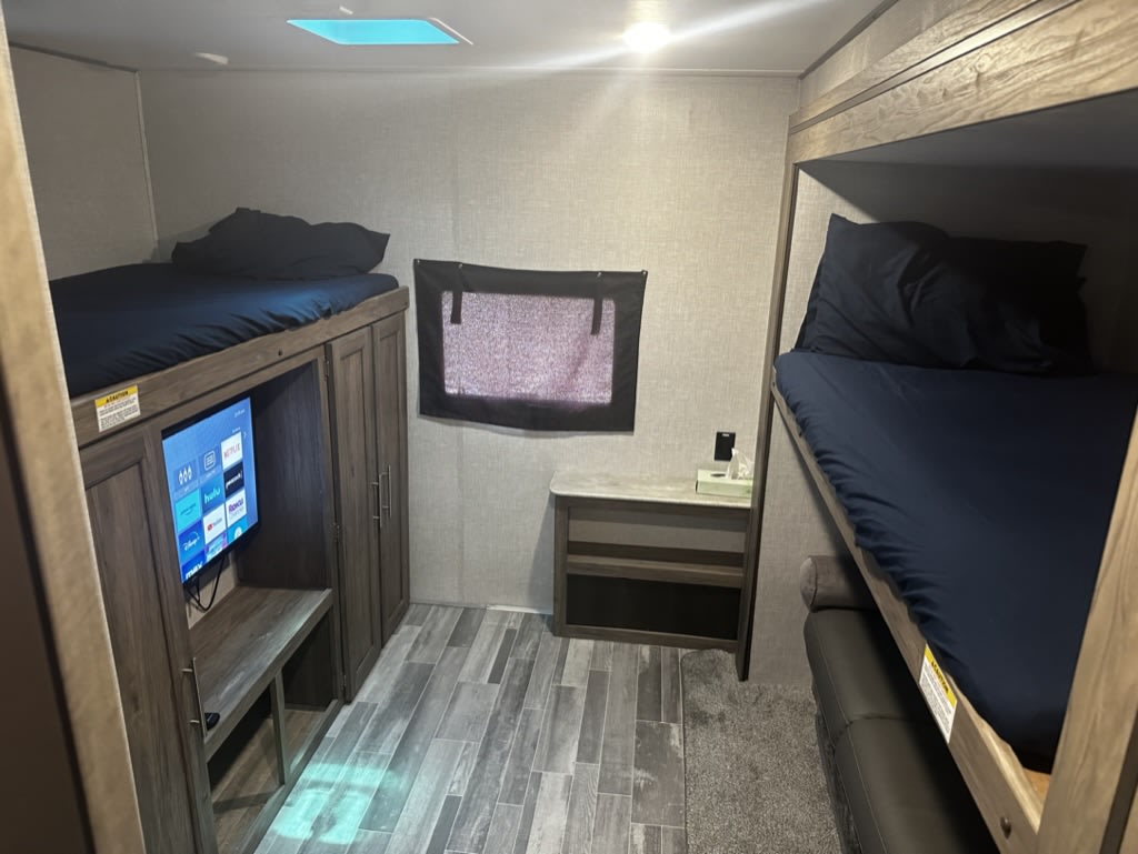 RV rental bunkhouse interior: Two sets of dark-sheeted bunk beds flank a TV and storage cabinet, offering cozy sleeping and entertainment.. Crossroads RV Zinger 2023