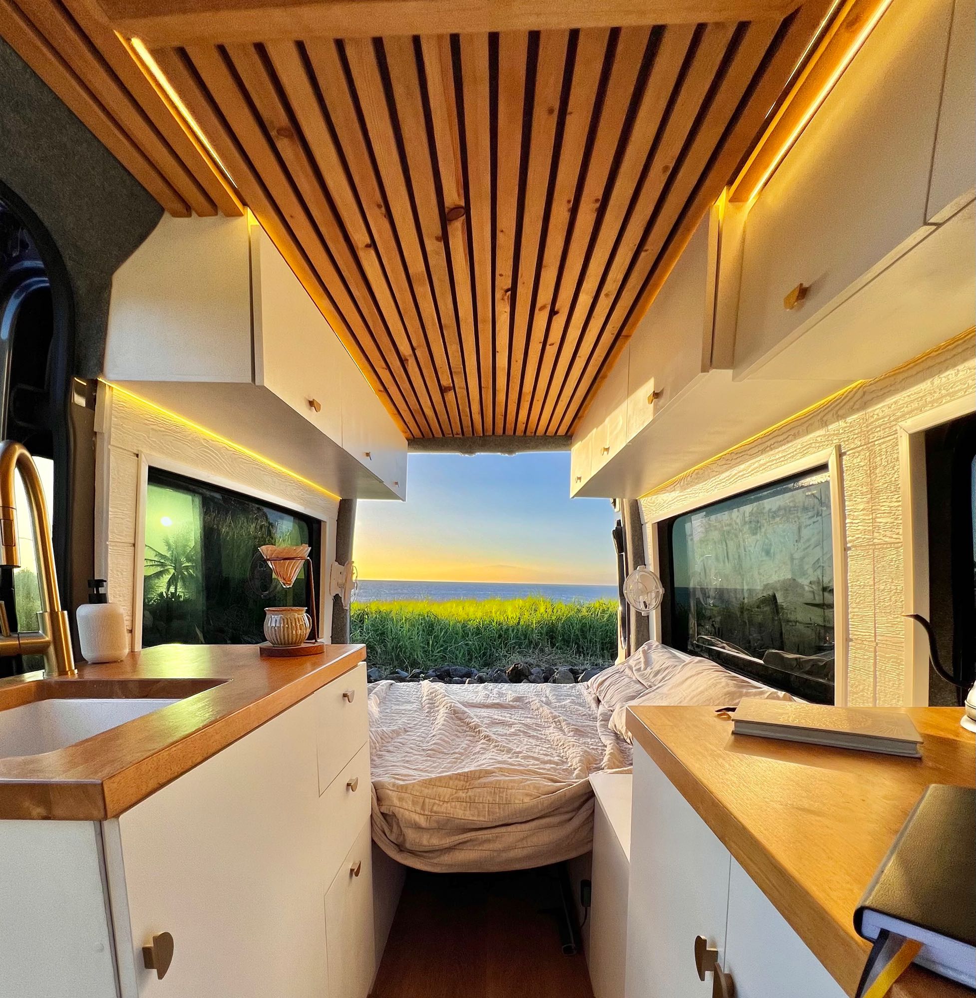 Inside this beautiful RV rental, a wooden ceiling and white interior frame a stunning ocean sunset view, offering ultimate van life comfort.. Mercedes-Benz Sprinter 2015