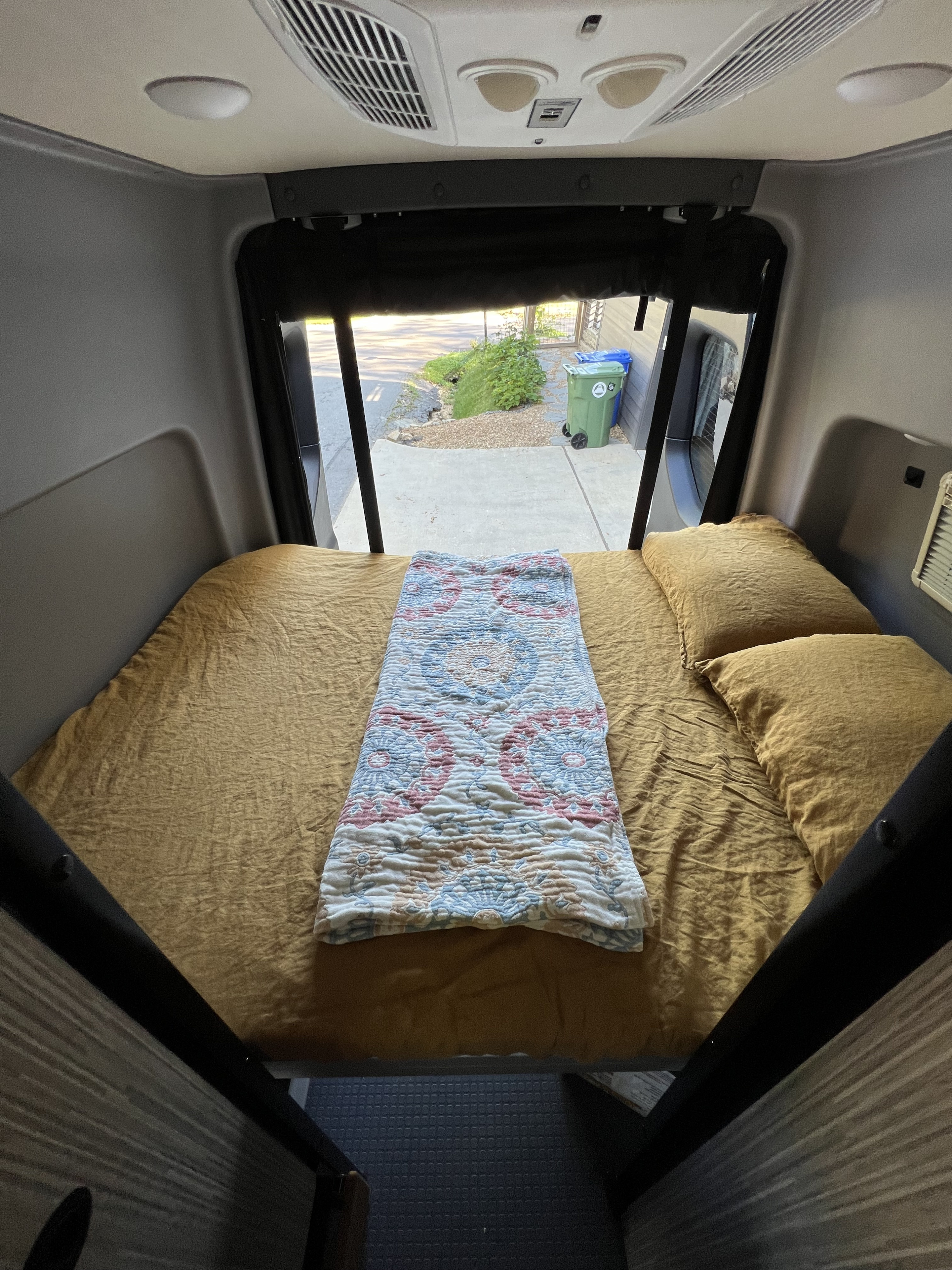 Cozy RV rental bed with mustard yellow sheets & patterned blanket inside a camper van. View out the back window.. Winnebago Revel 2023