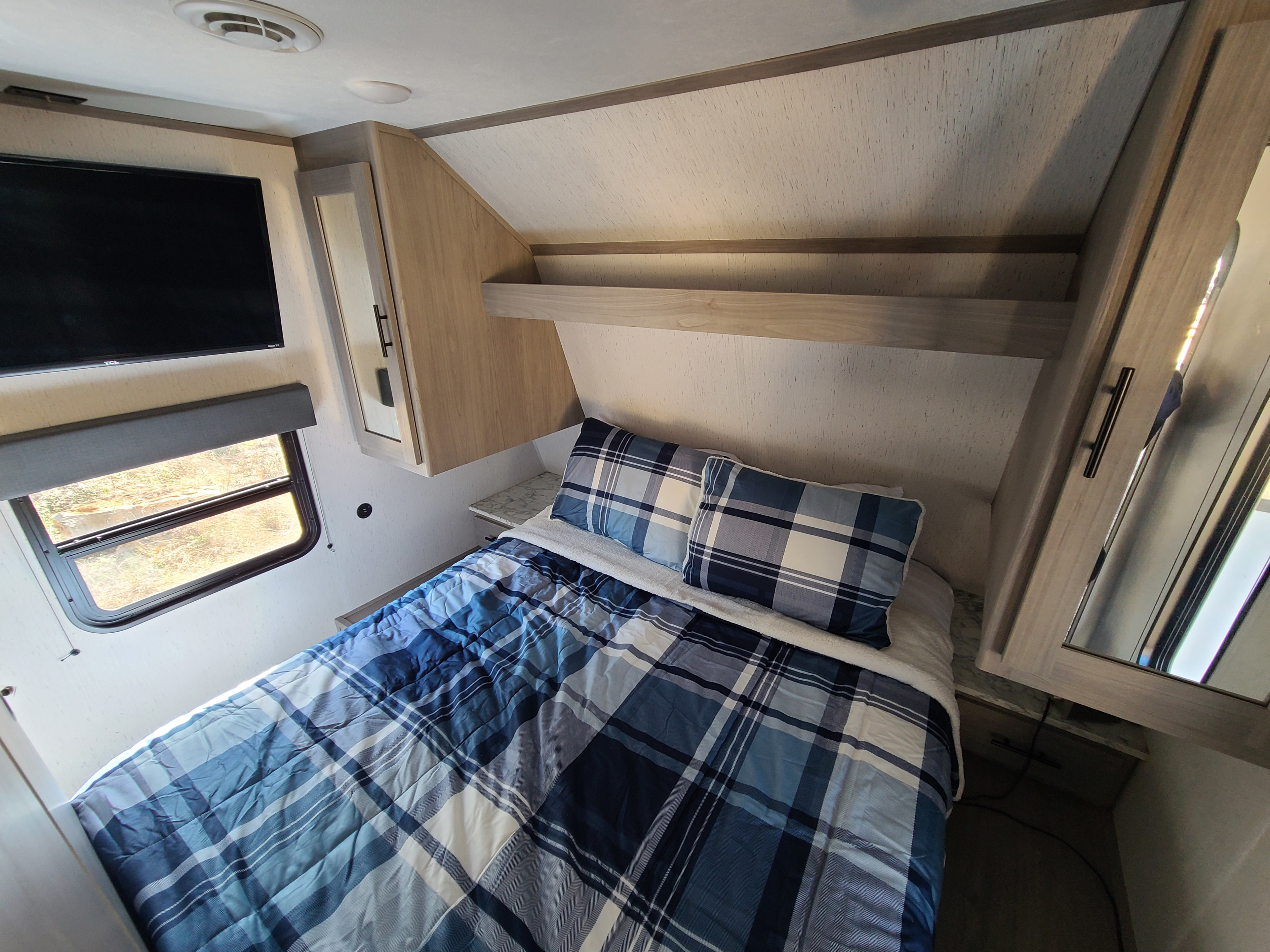 RV rental interior: A compact bedroom with a blue plaid bed, TV, window, and wood-toned cabinetry.. Dutchmen Coleman 2022