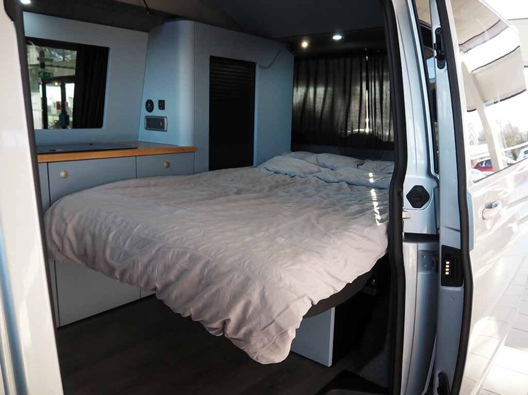 Inside an RV rental: a cozy bed with light grey bedding, a compact counter with integrated appliances and controls, and dark curtains.. Volkswagen T6 Transporter 2024