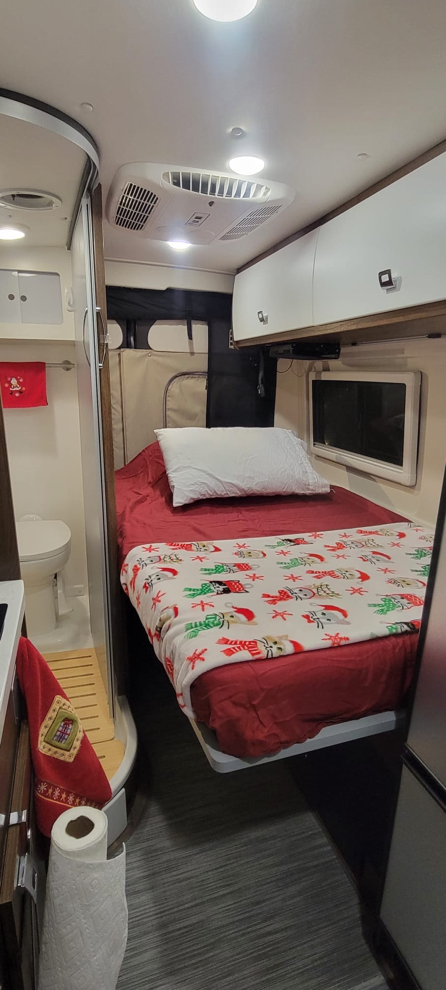 Inside a cozy rv rental, showing a bed with festive blanket, overhead storage, AC unit, and a compact bathroom with toilet.. Winnebago Travato 2022