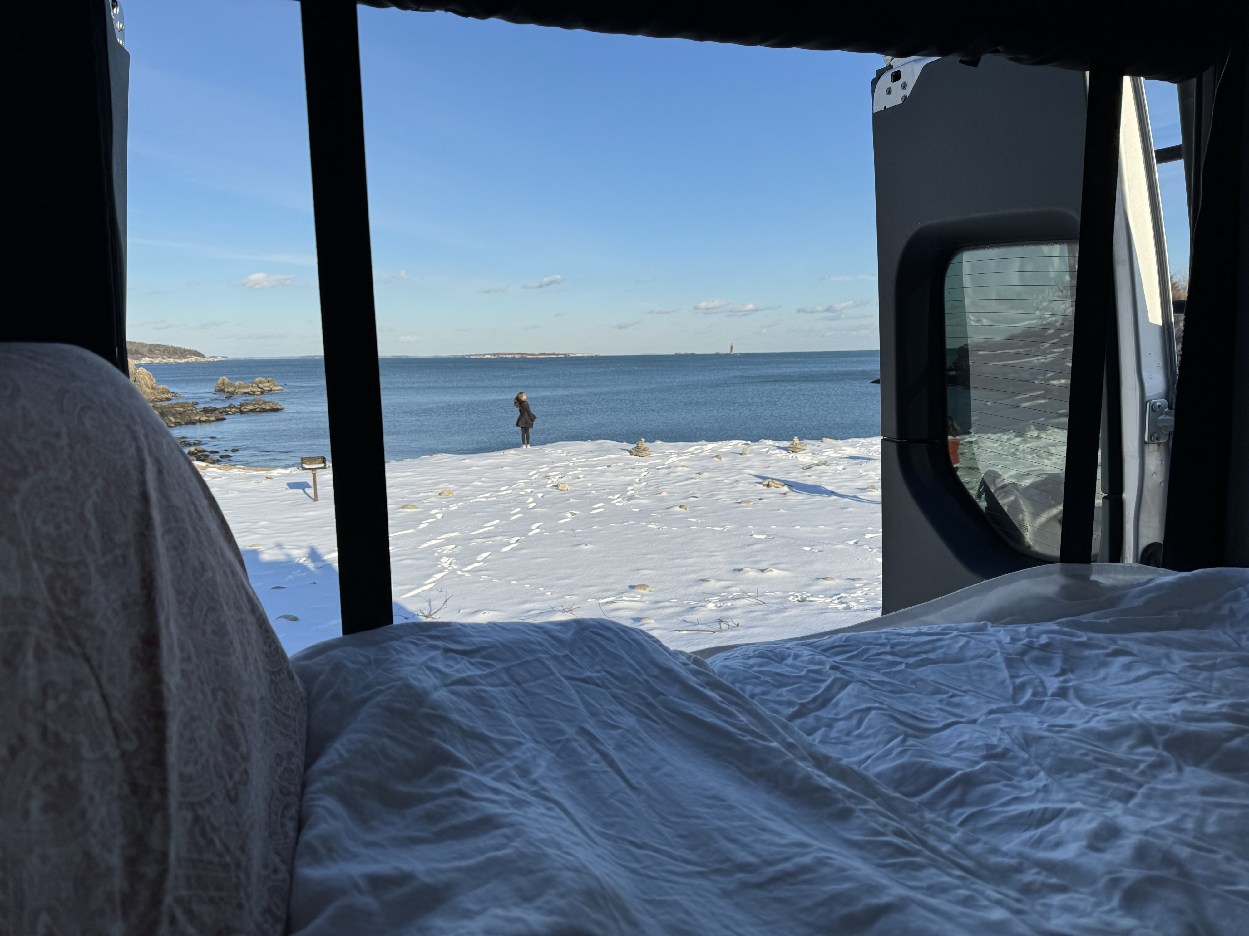 View from an RV rental's open back doors: a person on a snowy beach looks out at the ocean and distant lighthouse.. Winnebago Revel 2022