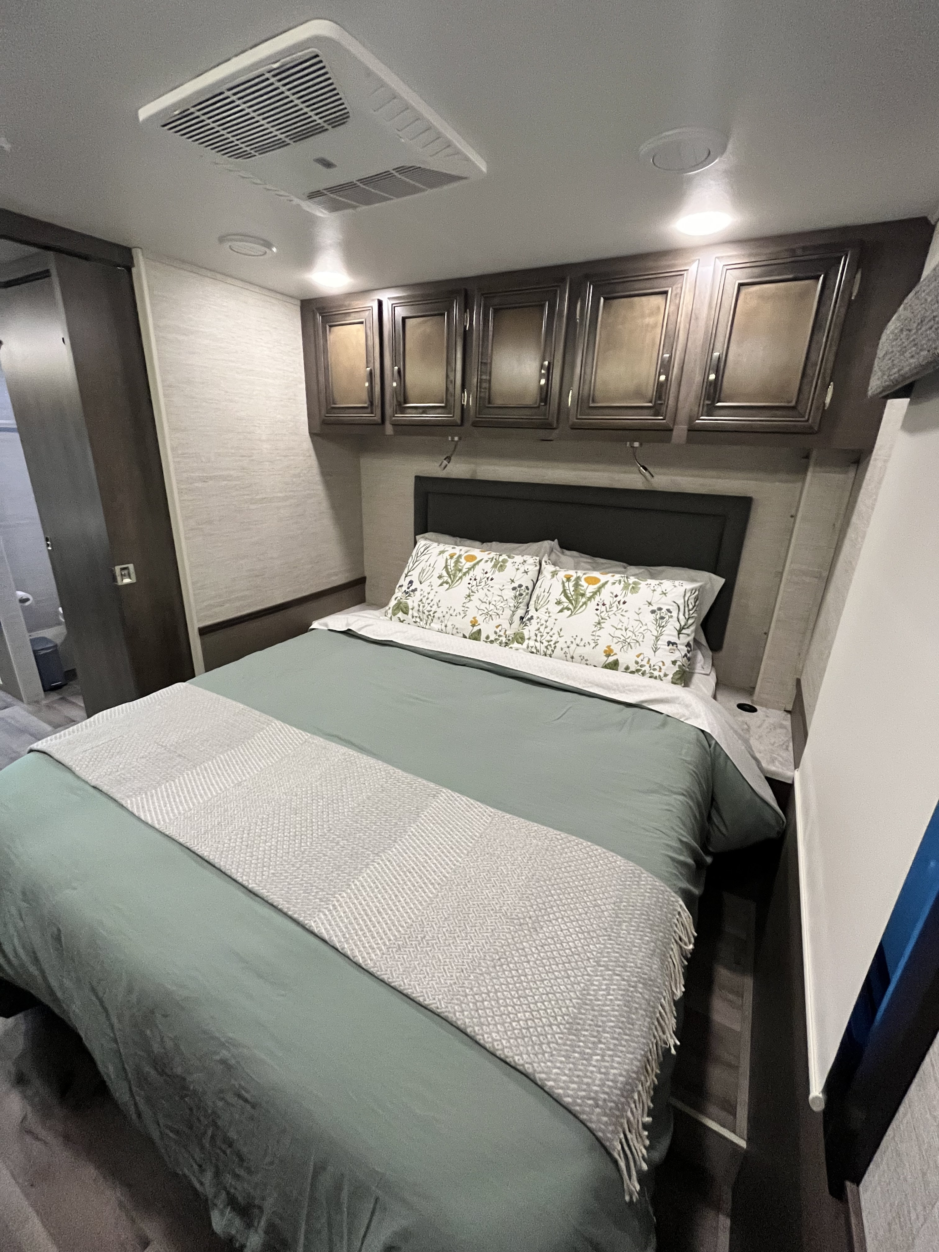 Cozy RV rental bedroom featuring a sage green bed with floral pillows, overhead cabinets, and ceiling-mounted AC unit.. Jayco Alante 2023