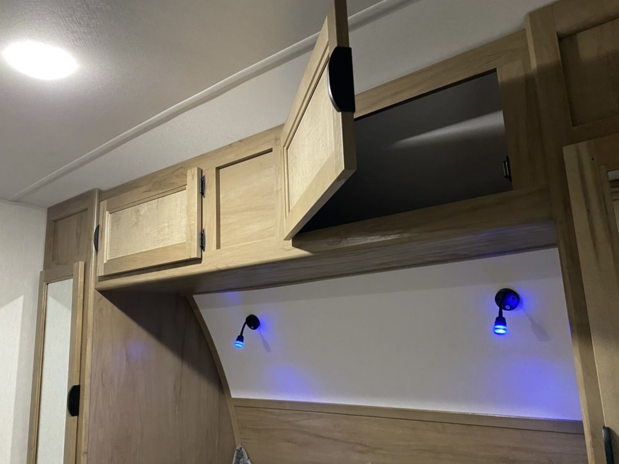 Cozy RV rental interior featuring light wood overhead cabinets, one open, and two blue LED reading lights above the bed.. Forest River Cherokee Grey Wolf 2024