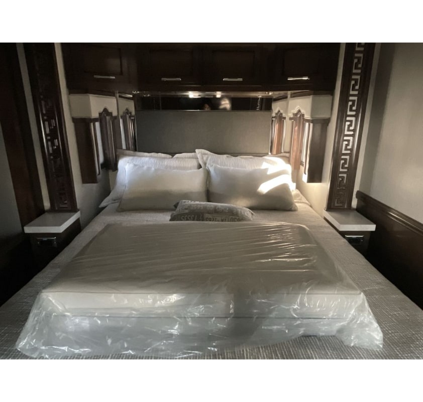 This RV rental bedroom features a bed with white pillows and a new, plastic-wrapped mattress topper, surrounded by dark wood cabinetry.. Newmar Ventana 2023