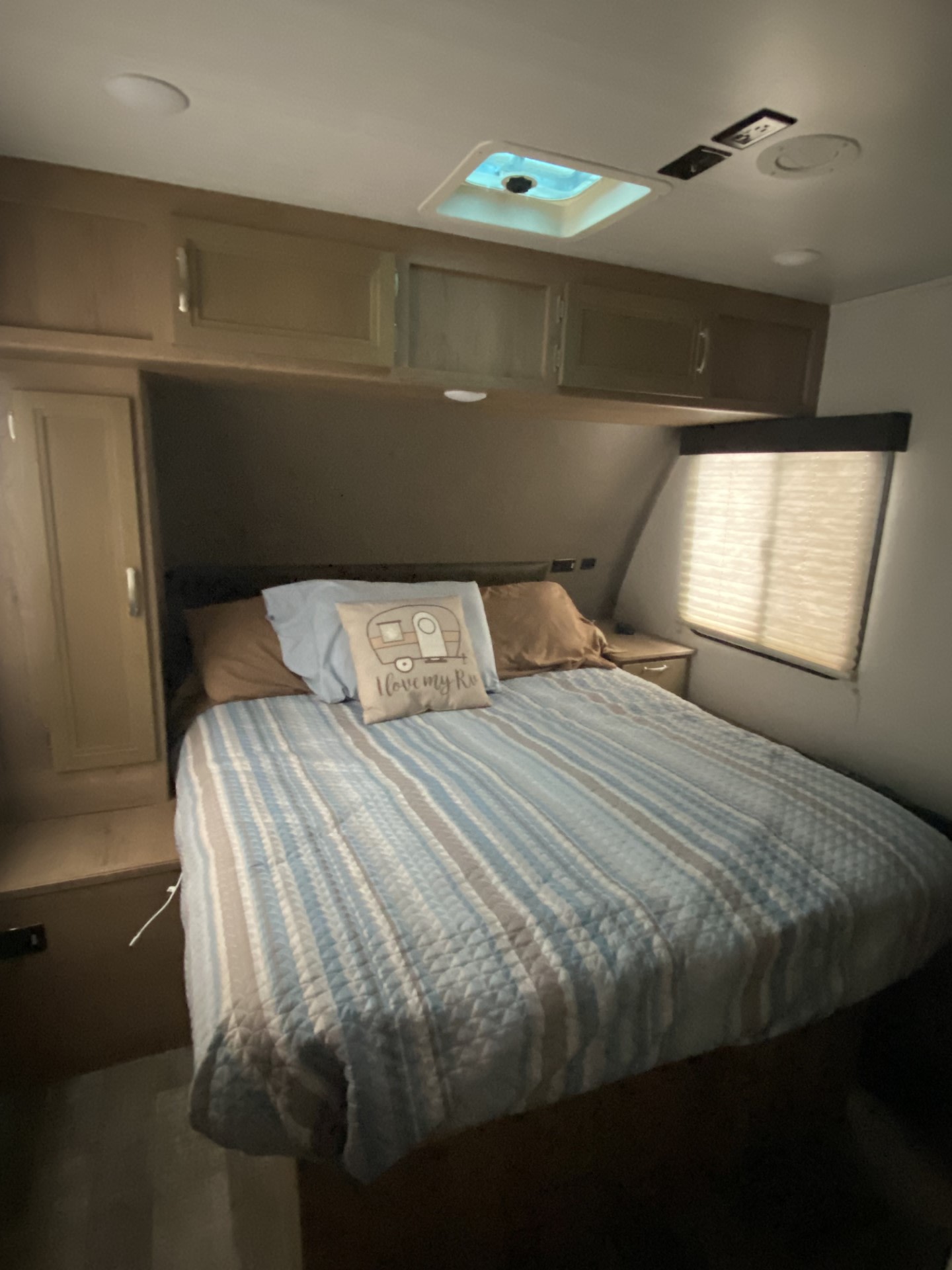 Cozy RV rental bedroom with blue striped bedding, "I love my RV" pillow, overhead cabinets, and window with blinds.. Winnebago Minnie Winnie 2020