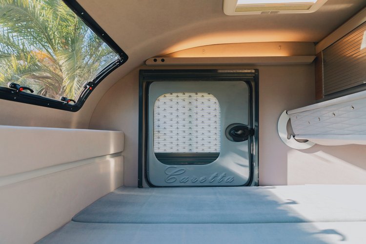 Inside a cozy rv rental: a comfy bed, window with palm trees, "Caretta" door, and an upper bunk/shelf.. Caretta + NaïtUp Quättro 1500 + Quättro 2024