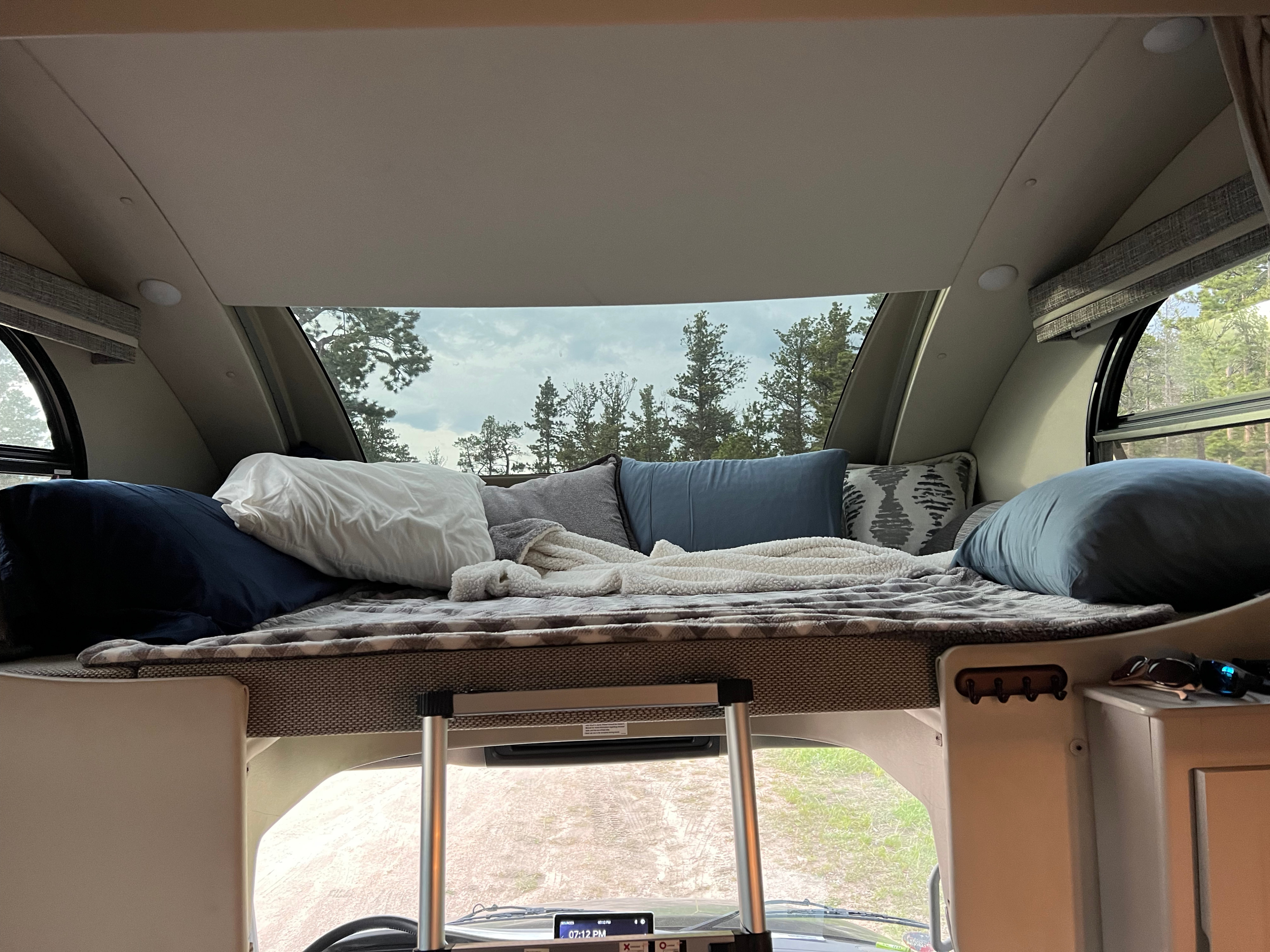 Cozy RV rental bunk bed with plush pillows and blankets, offering a peaceful forest view at 7:12 PM.. Thor Inception 2023