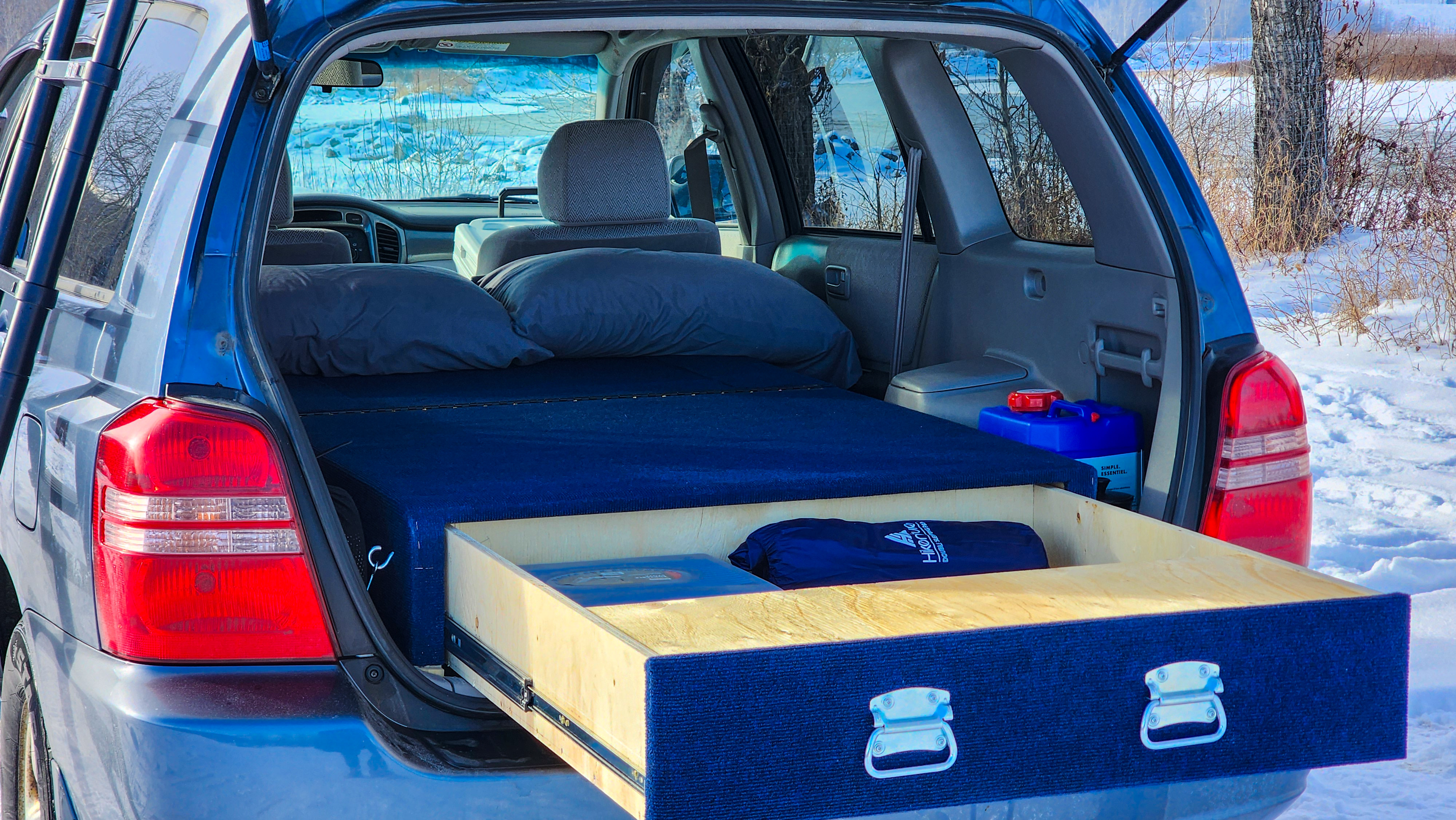 Inside a blue SUV, a custom bed and a pulled-out wooden drawer with items create a car camping setup, a versatile RV rental option.. Toyota Highlander 2003
