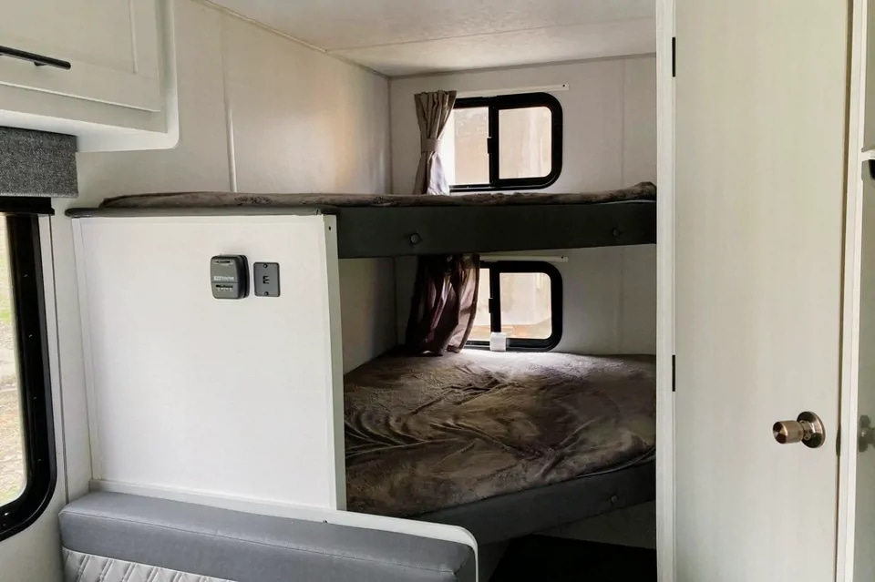 Inside an RV rental: view of bright bunk beds with windows and curtains. Wall-mounted controls are visible near the lower bunk.. Forest River Salem FSX 2021