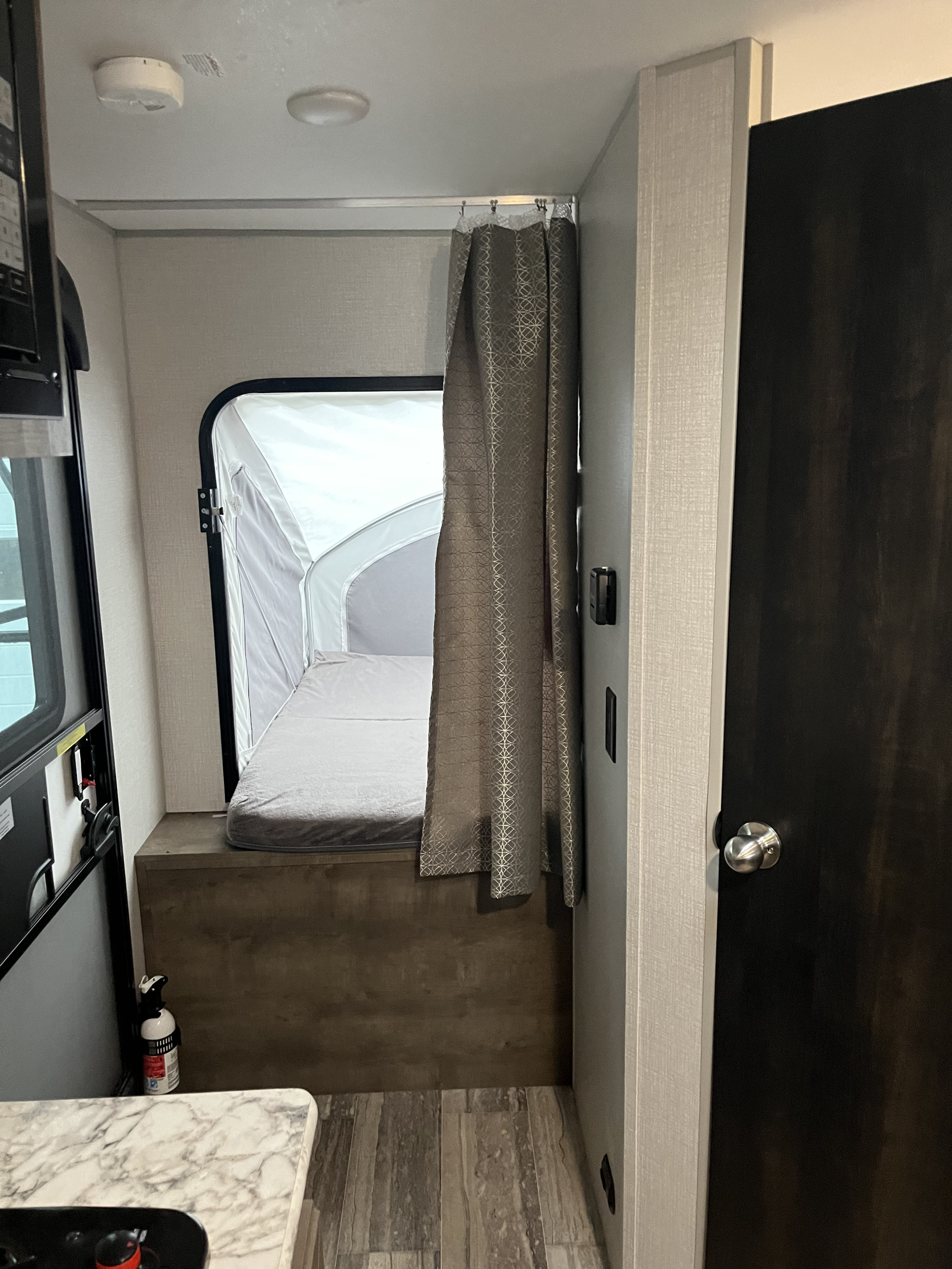A clean RV rental interior featuring a pop-out bed section, privacy curtain, and adjacent kitchen counter. Essential safety devices visible.. KZ sportsman 2021