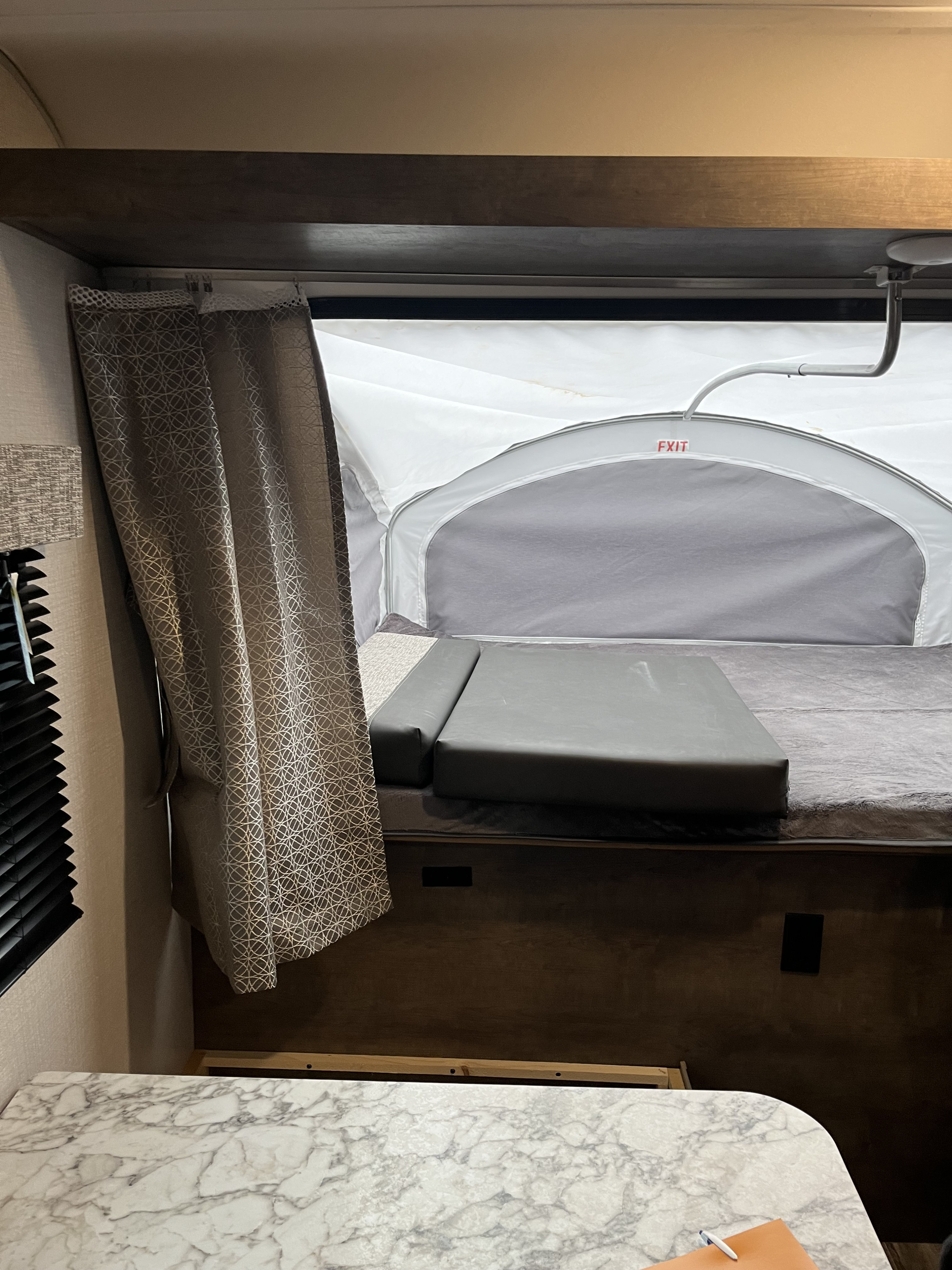 RV rental interior: Pop-up camper bed, patterned curtain, and table. The expandable tent section includes an 'EXIT' sign.. KZ sportsman 2021