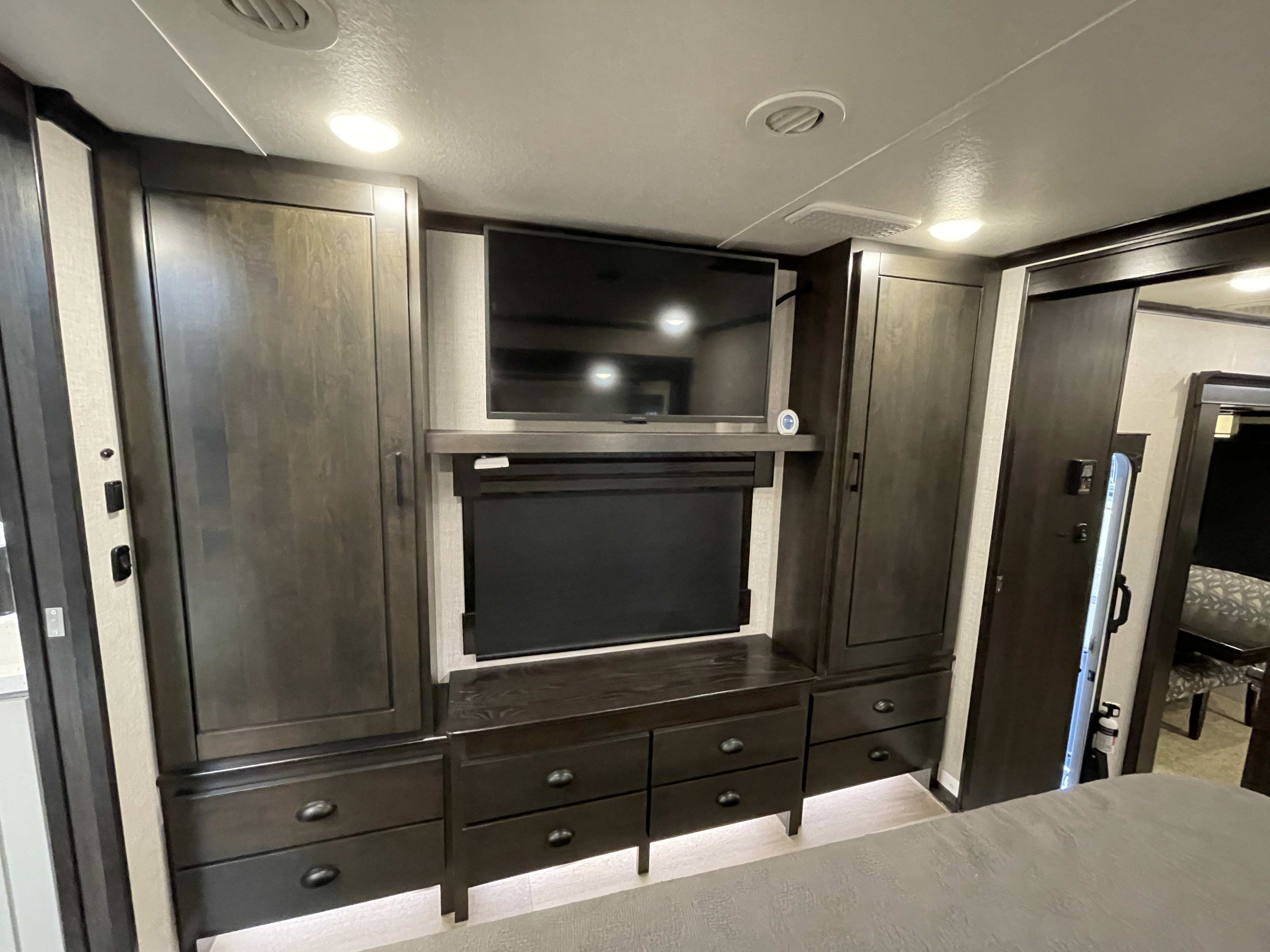 Modern RV rental interior showing a dark wood bedroom wall unit with TV, storage, and sliding door. Ambient lighting adds comfort.. Crossroads Redwood 2021