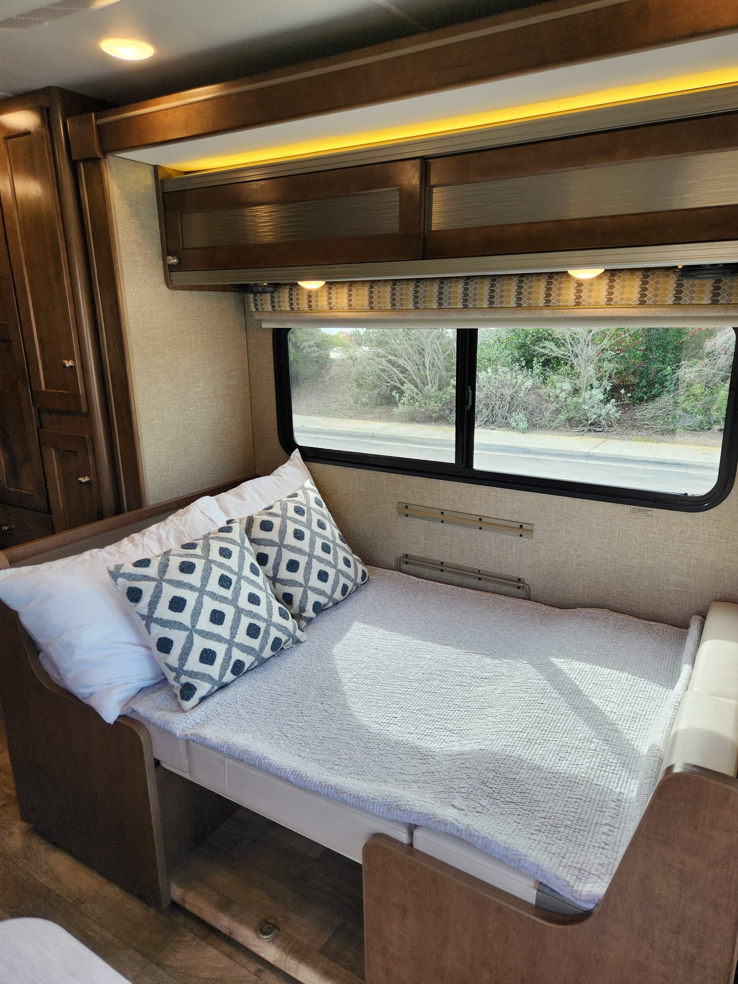 Spacious RV rental interior featuring a bed with patterned pillows, overhead cabinets, and a scenic window view.. Winnebago Fuse 2019
