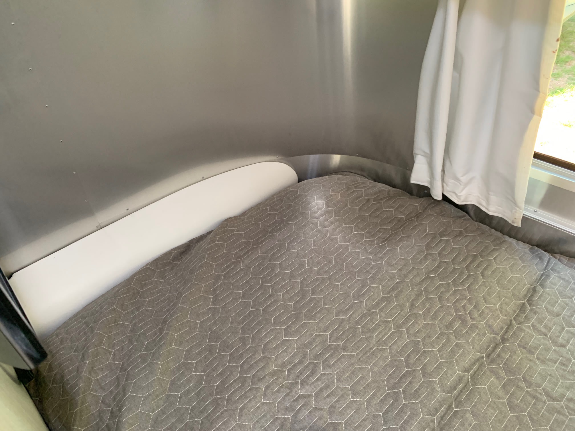 Angled view of a compact RV rental bed with a gray hexagonal quilted duvet, white curved panel, and polished aluminum walls.. Airstream Sport 2019