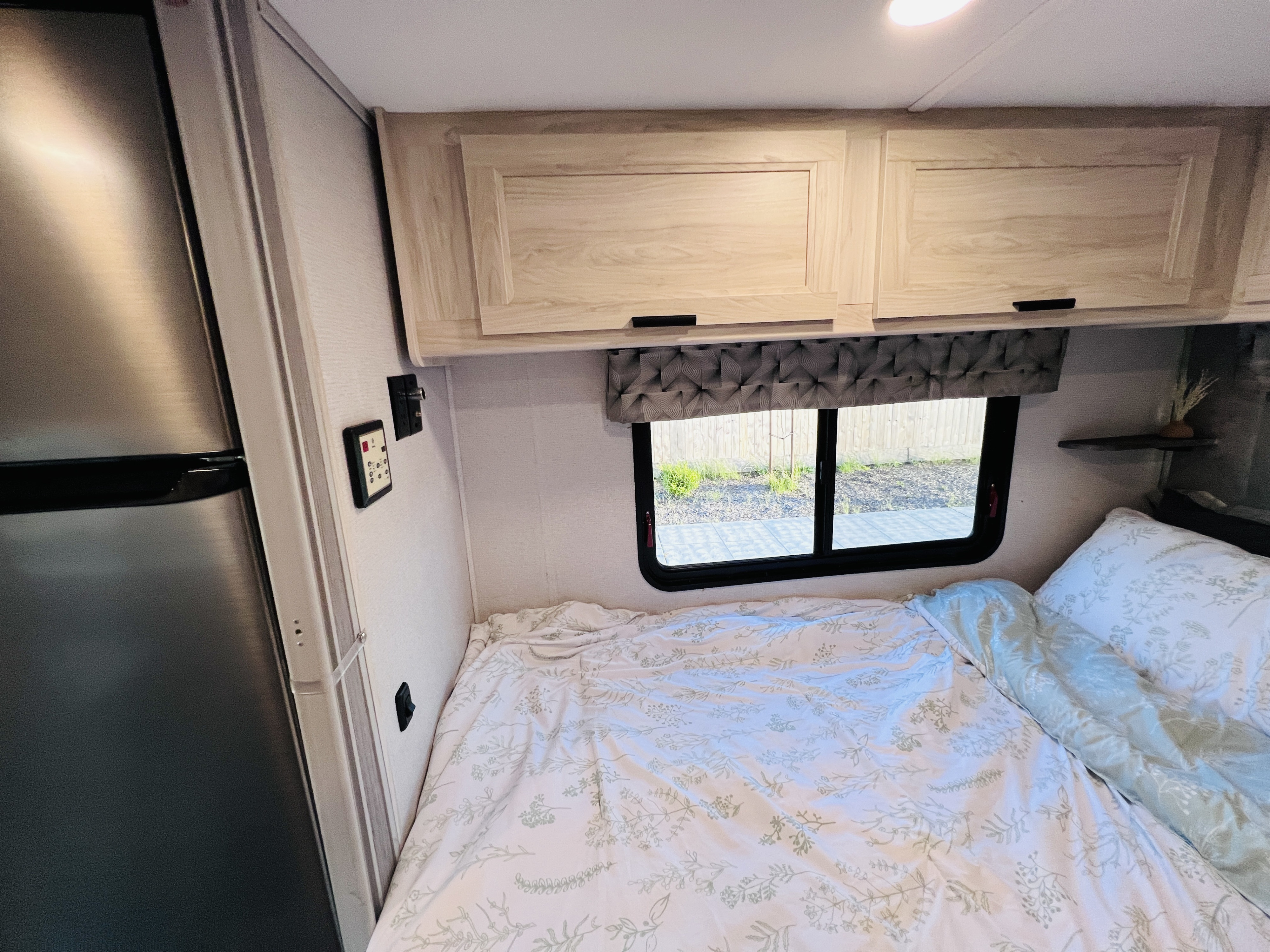 This rv rental features a cozy bedroom with a bed, light wood overhead cabinets, window, and a built-in stainless steel fridge.. Forest River Sunseeker LE 2022