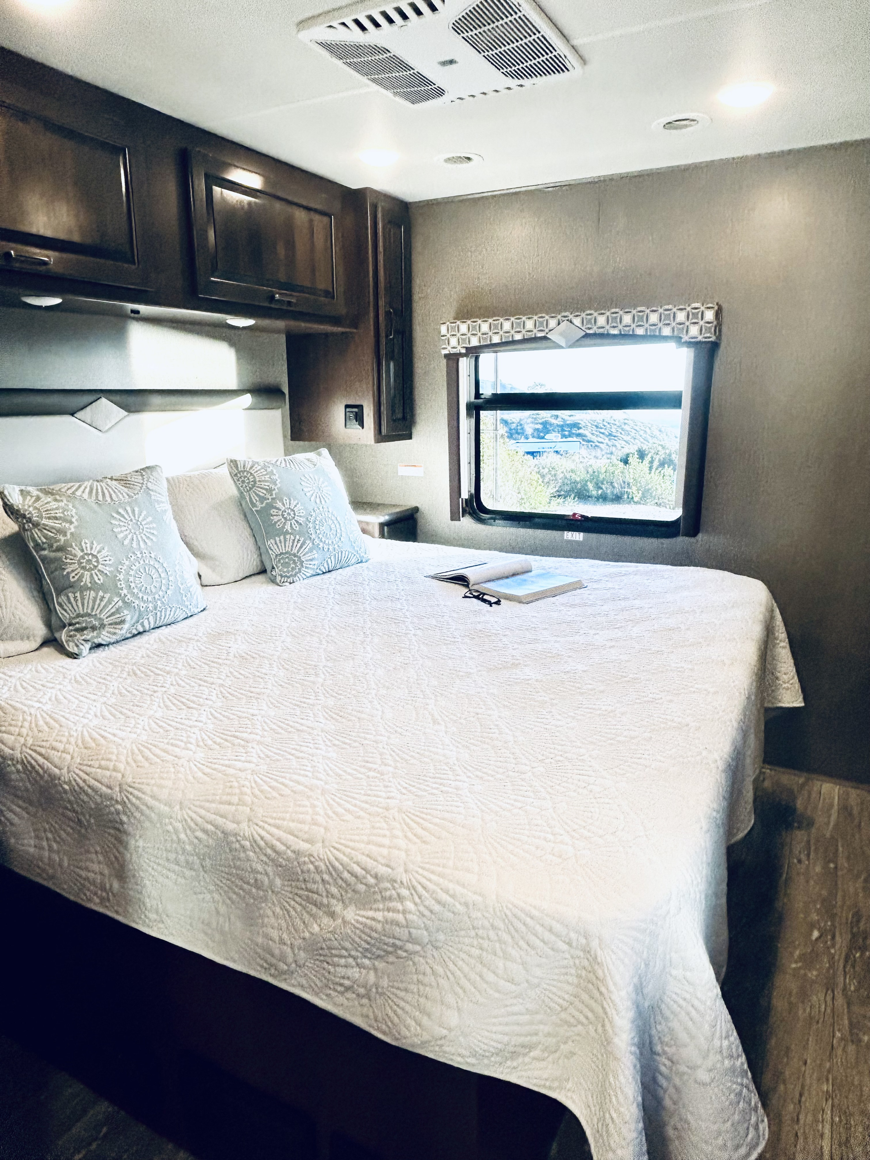Cozy RV rental bedroom featuring a comfortable white bed with patterned pillows, overhead cabinets, and a window with a scenic view.. Thor Motor Coach Hurricane 2020
