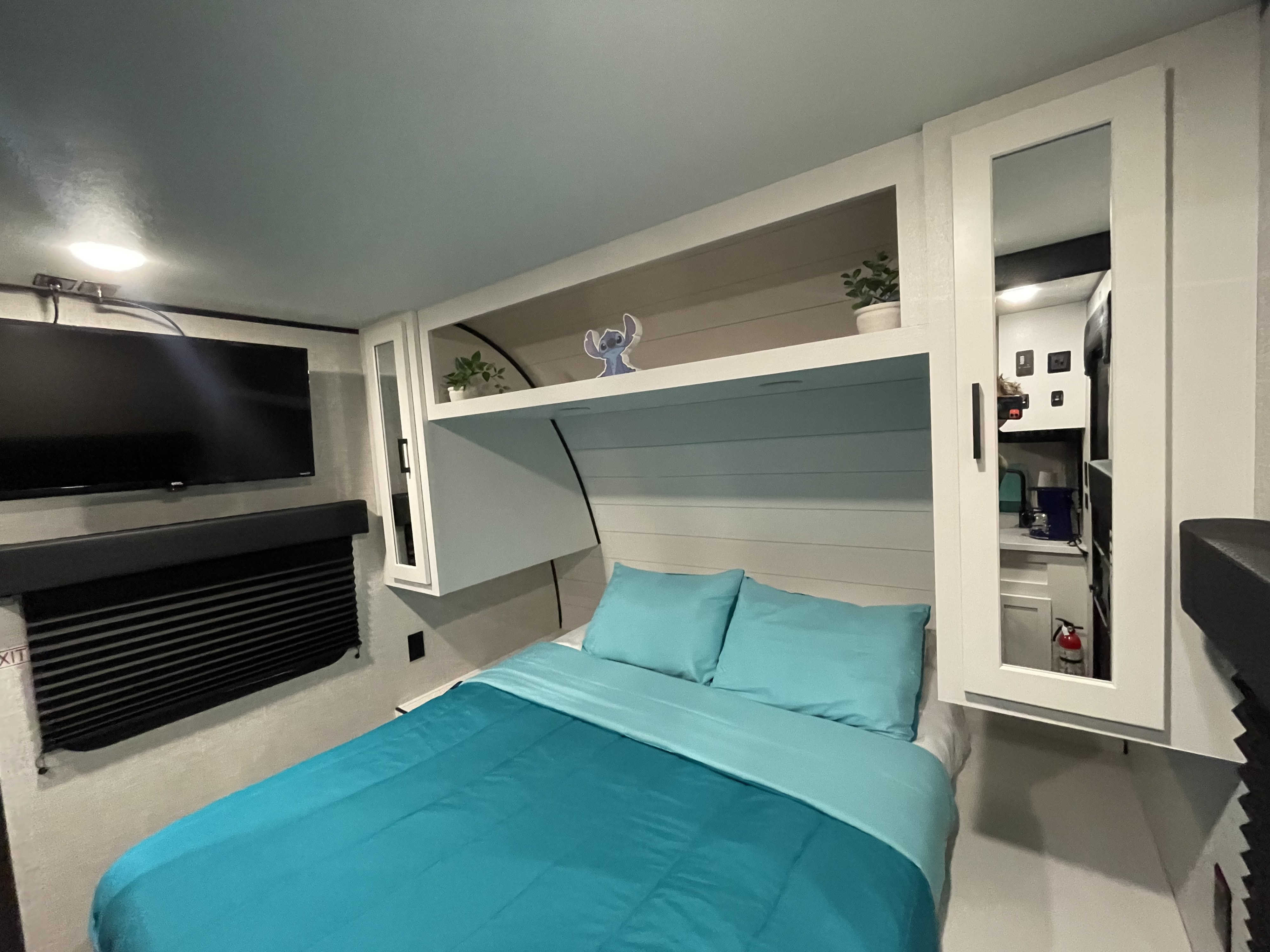 Step into this modern RV rental bedroom, featuring a comfy teal bed, TV, clever overhead storage, and a reflected kitchen view.. Jayco Jay Flight 261BHS 2024
