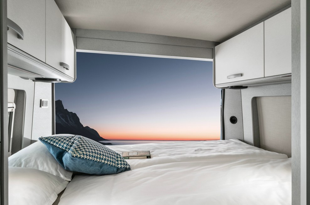 Inside a comfortable RV rental bedroom, a pristine bed faces a breathtaking mountain and sunset vista.. Hymer FREE 600 CAMPUS 2024