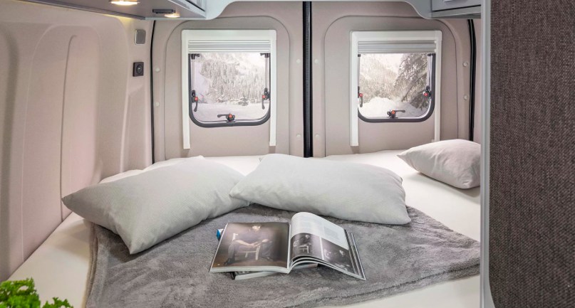 Relax in this RV rental bedroom. A cozy bed with pillows and a magazine, looking out two windows at a snowy mountain landscape.. Weinsberg Caratour Ford 550 MQ 2024