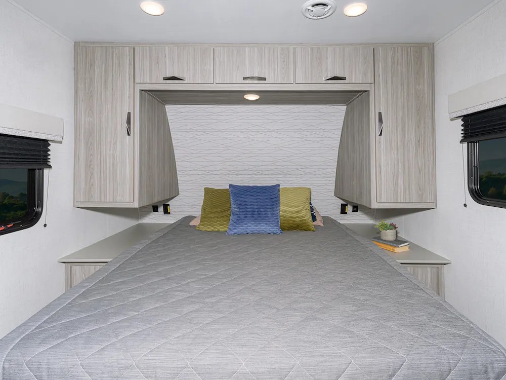 This RV rental bedroom features light wood cabinets, a comfy bed with three pillows, and windows. A bright and functional space.. Keystone RV 229bh/we 2024
