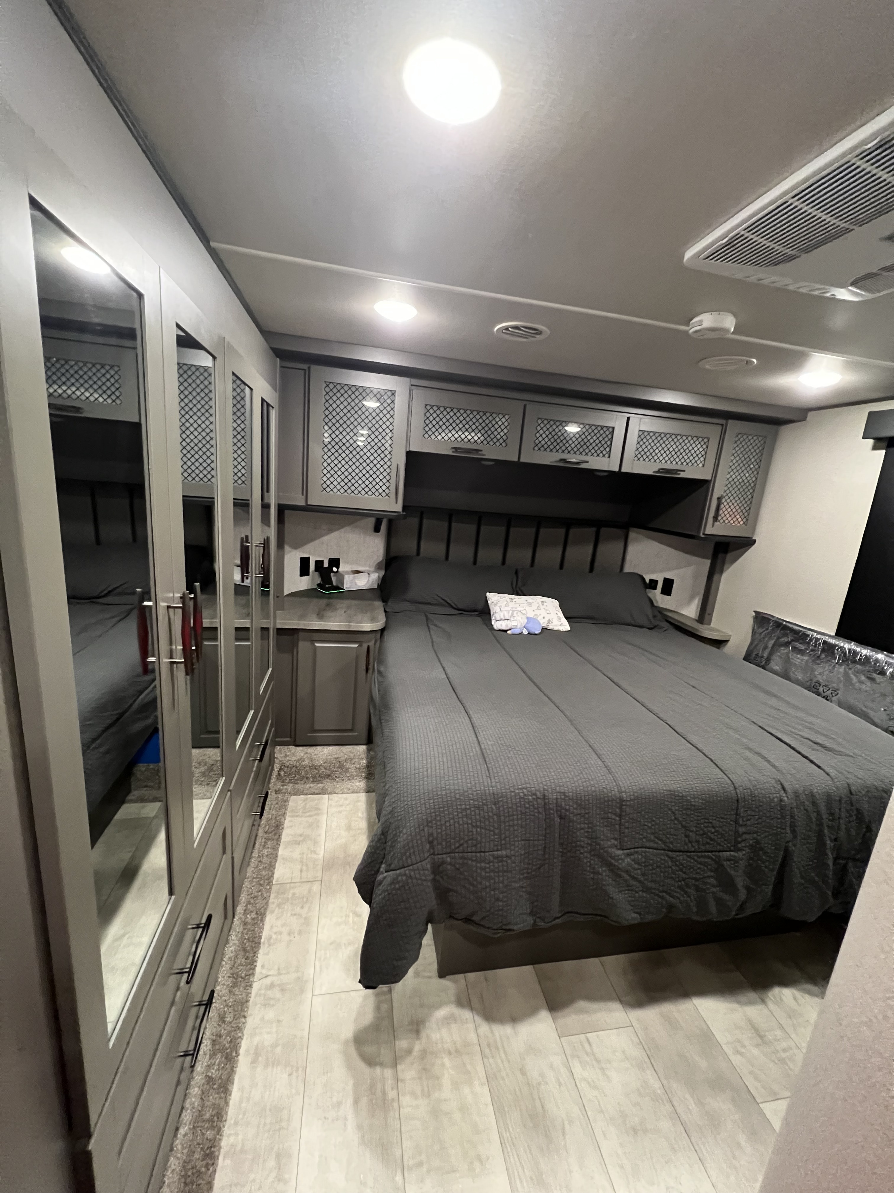 This RV rental bedroom offers a comfortable grey bed, built-in nightstands, overhead cabinets, and mirrored wardrobes for storage.. Grand Design Momentum 2021