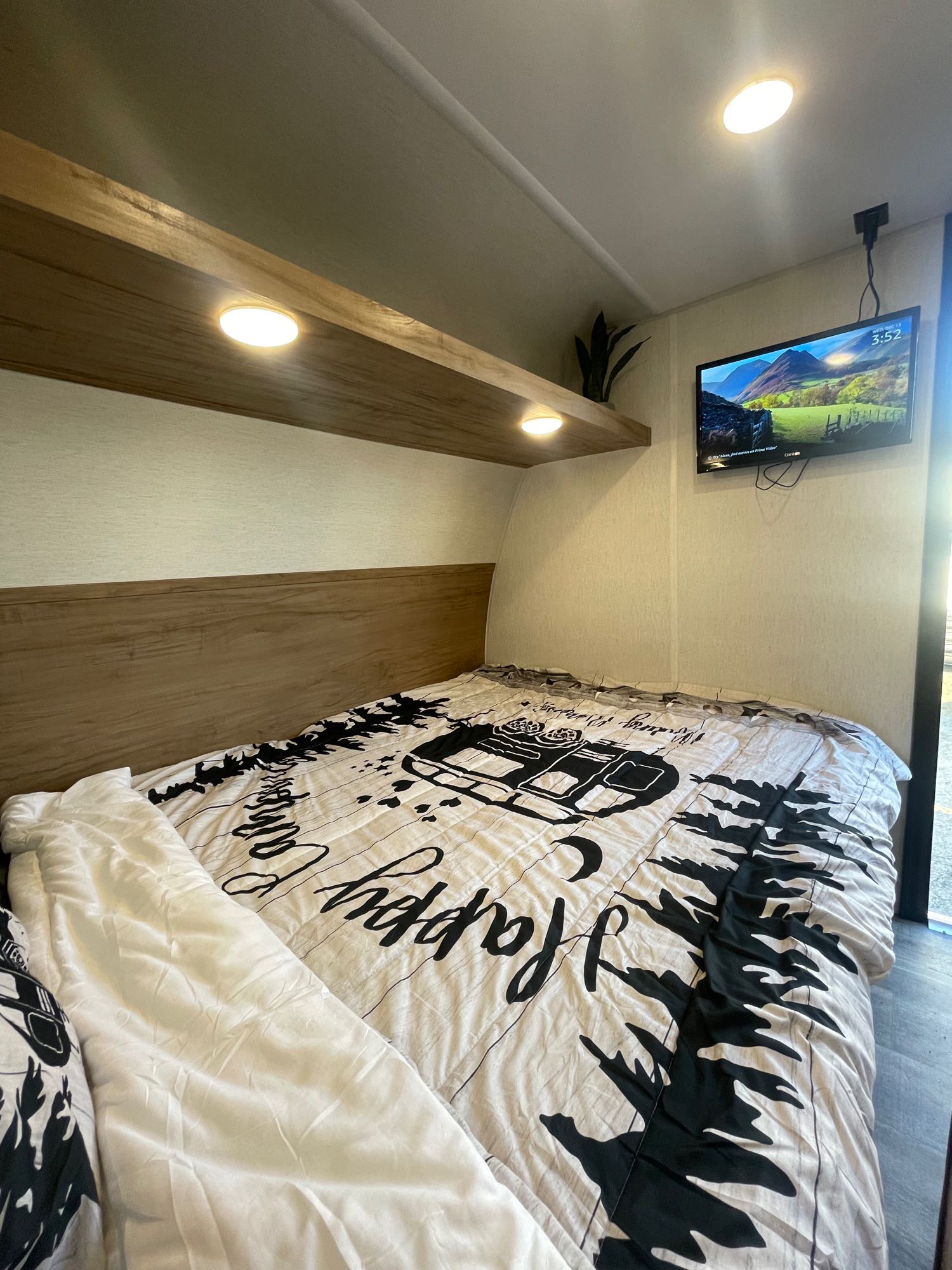 Inside a comfortable rv rental, featuring a cozy bedroom with a patterned duvet, wall-mounted TV, and warm shelf lighting.. Forest River Wolf Den 16EV 2024