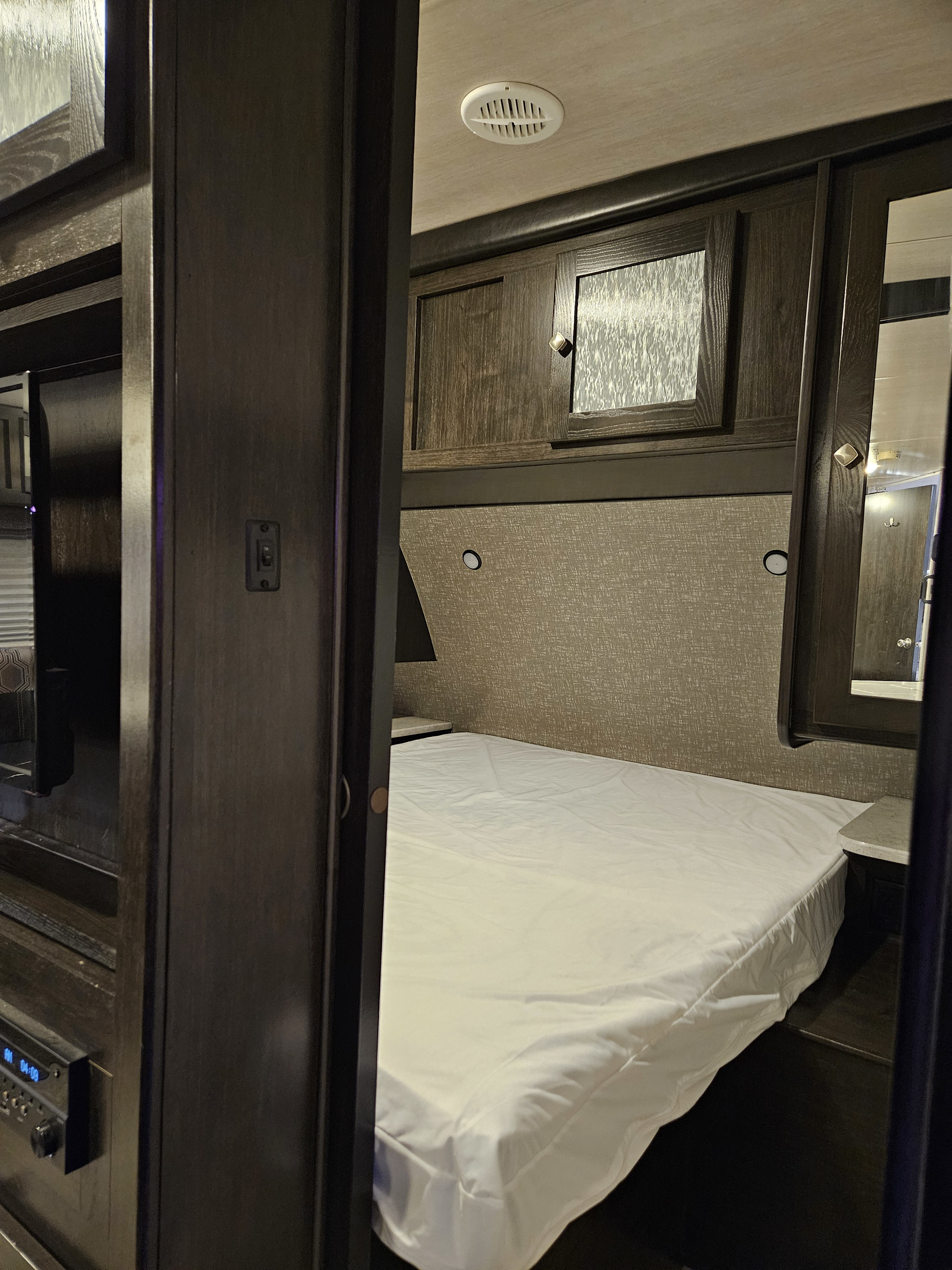 Compact RV rental bedroom with a white bed, dark wood overhead cabinets, a nightstand, and an entertainment system.. Heartland RVs North Trail 2019