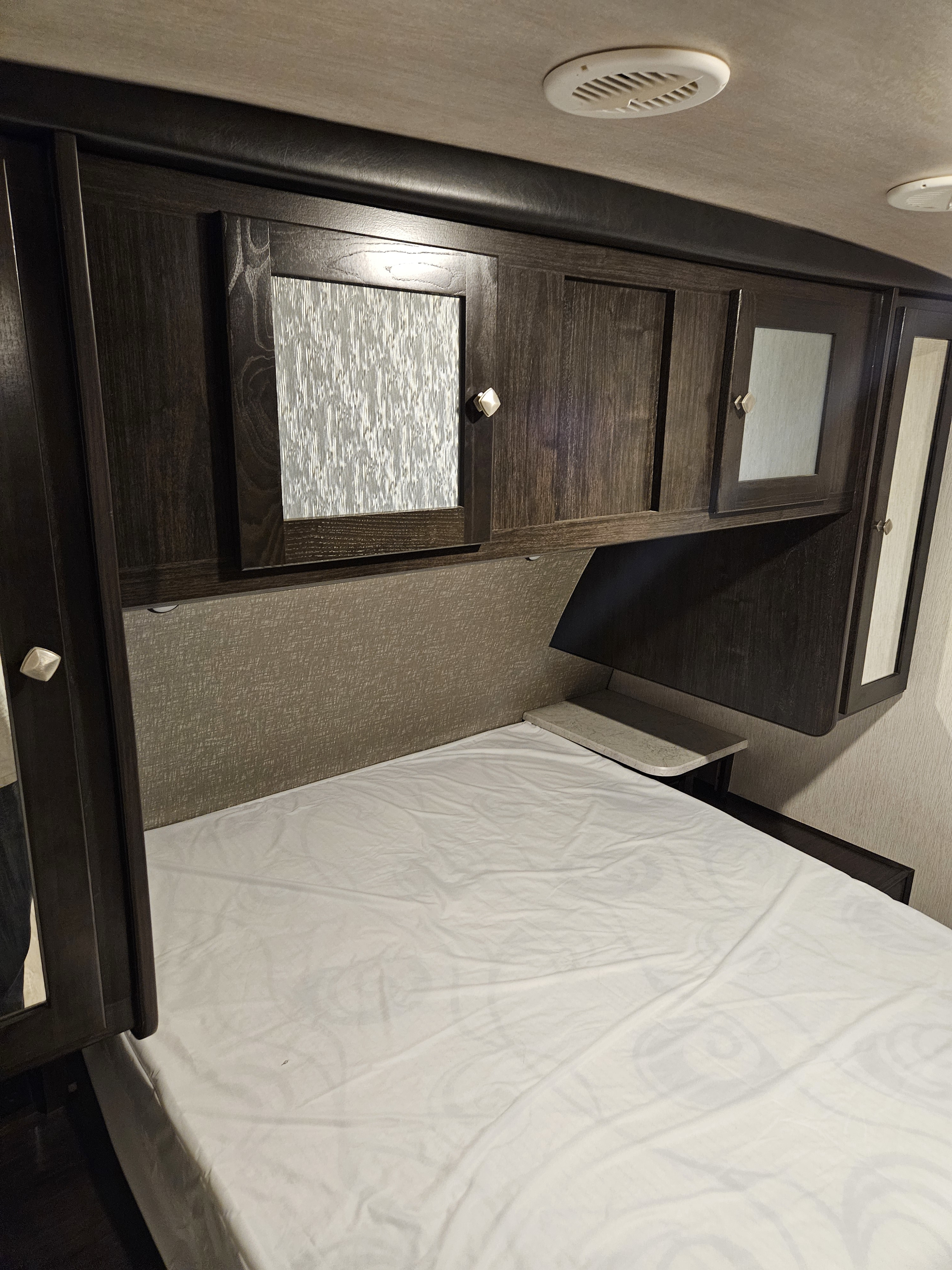 An RV rental bedroom with dark wood cabinets, a bed with white sheets, and ceiling vents, showing compact, functional design.. Heartland RVs North Trail 2019