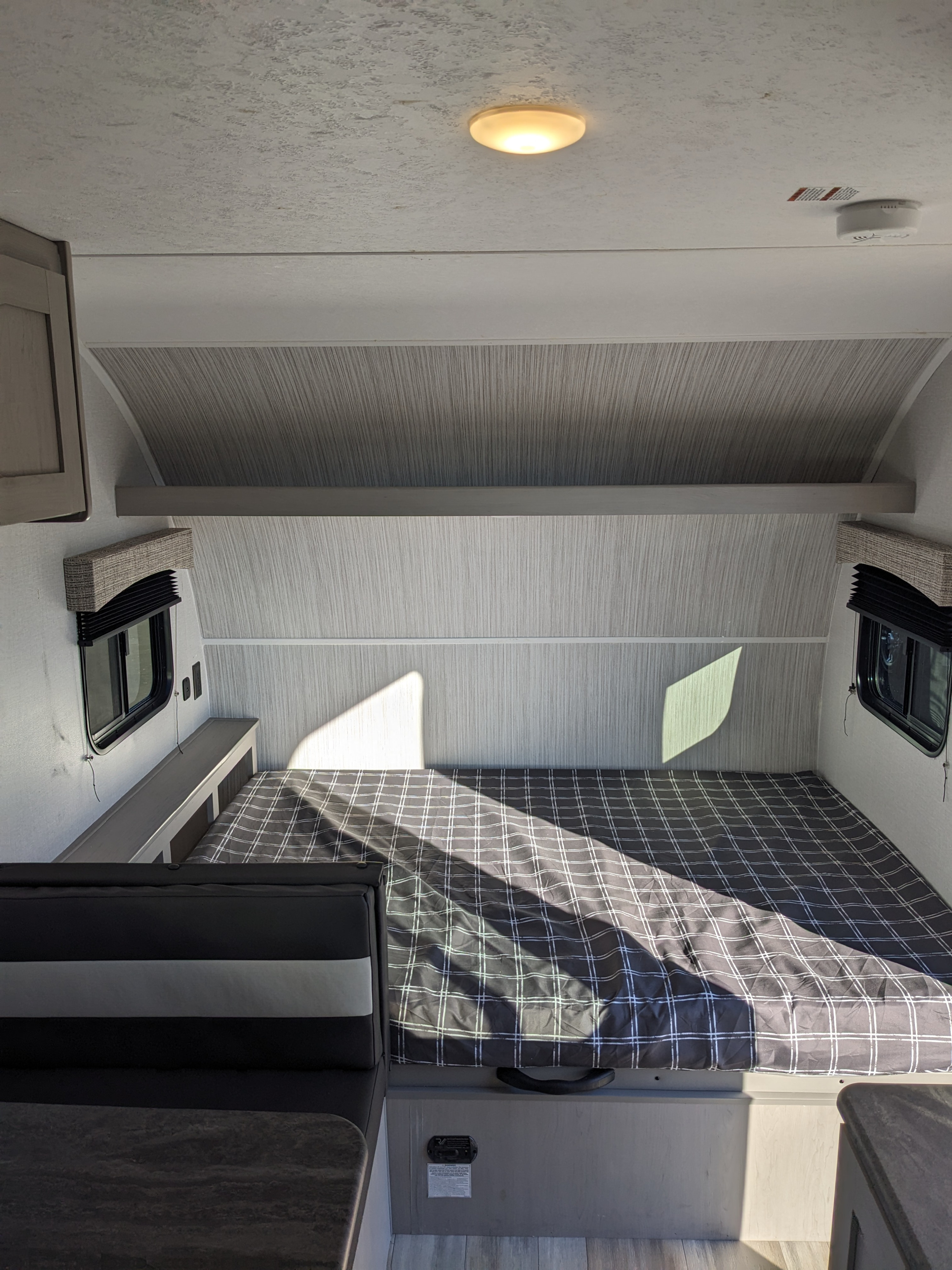 Interior view of an RV rental bedroom featuring a plaid bed, side windows with blinds, and an attached dining area.. Forest River Viking 3K 2023