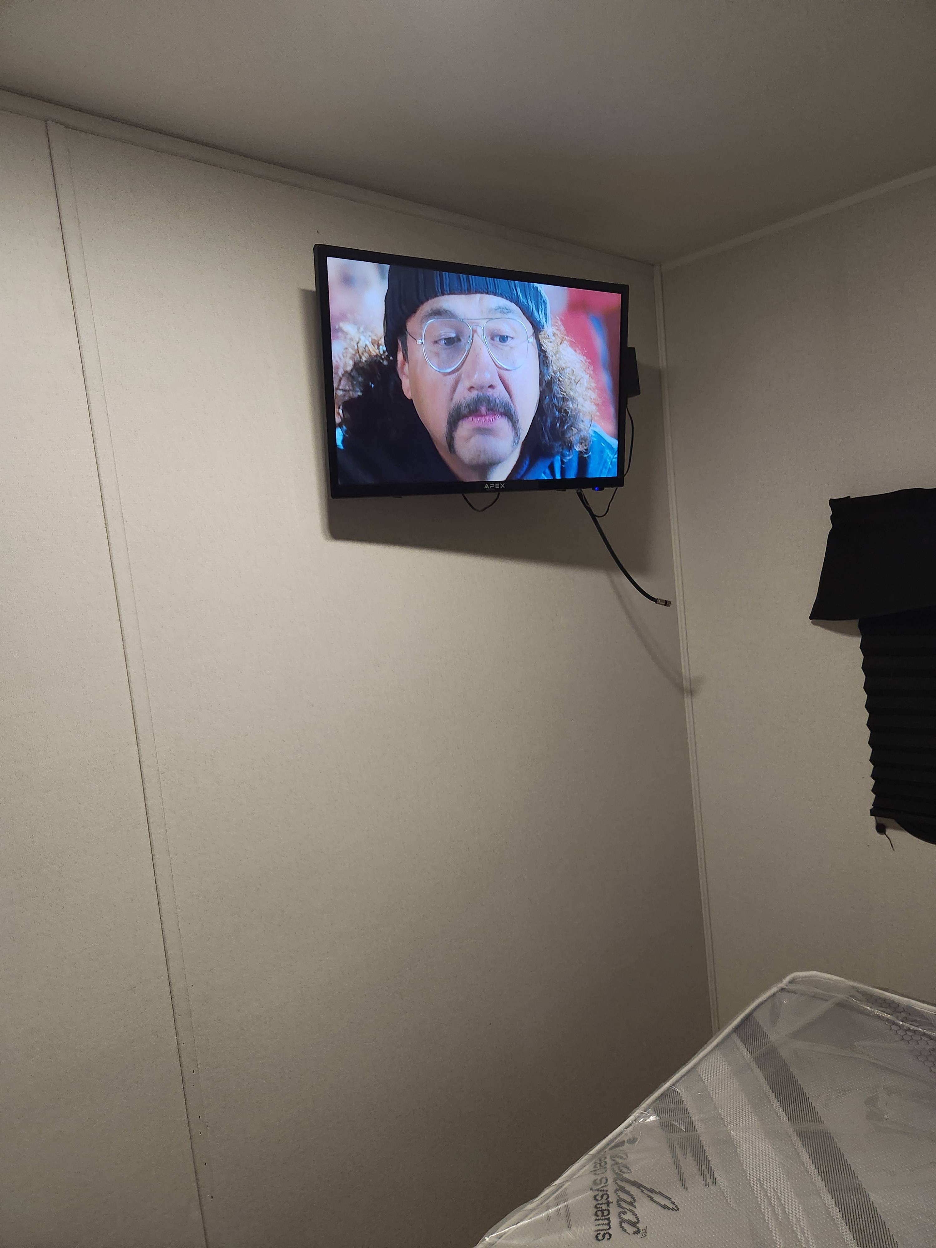 Inside an RV rental: A wall-mounted TV shows a man with a mustache and glasses. A new mattress is wrapped in plastic.. Heartland RVs Pioneer 2024