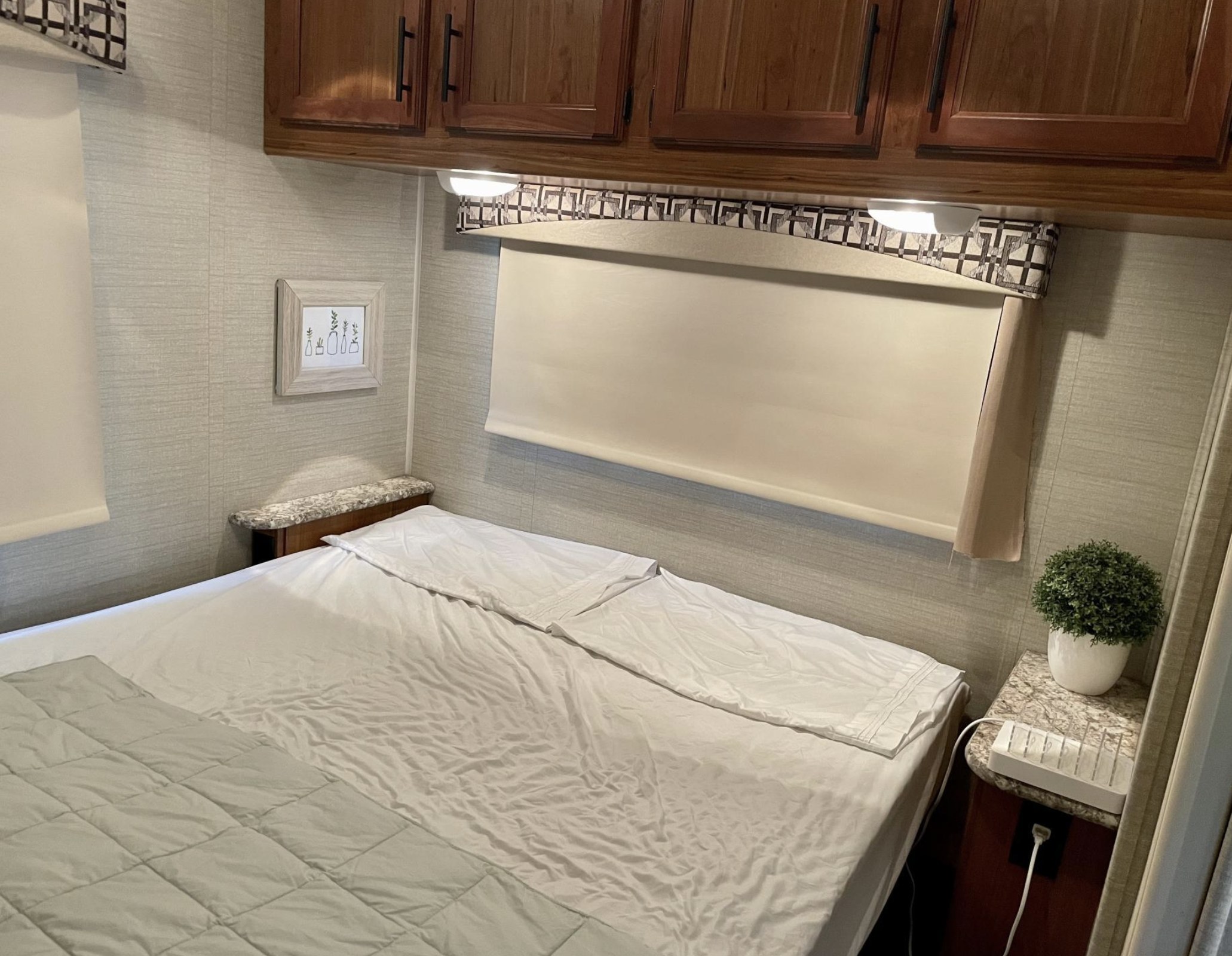 Cozy bedroom in an RV rental featuring a bed, window with blind, overhead cabinets, and small nightstands with decor and a charger.. Jayco Redhawk 2017