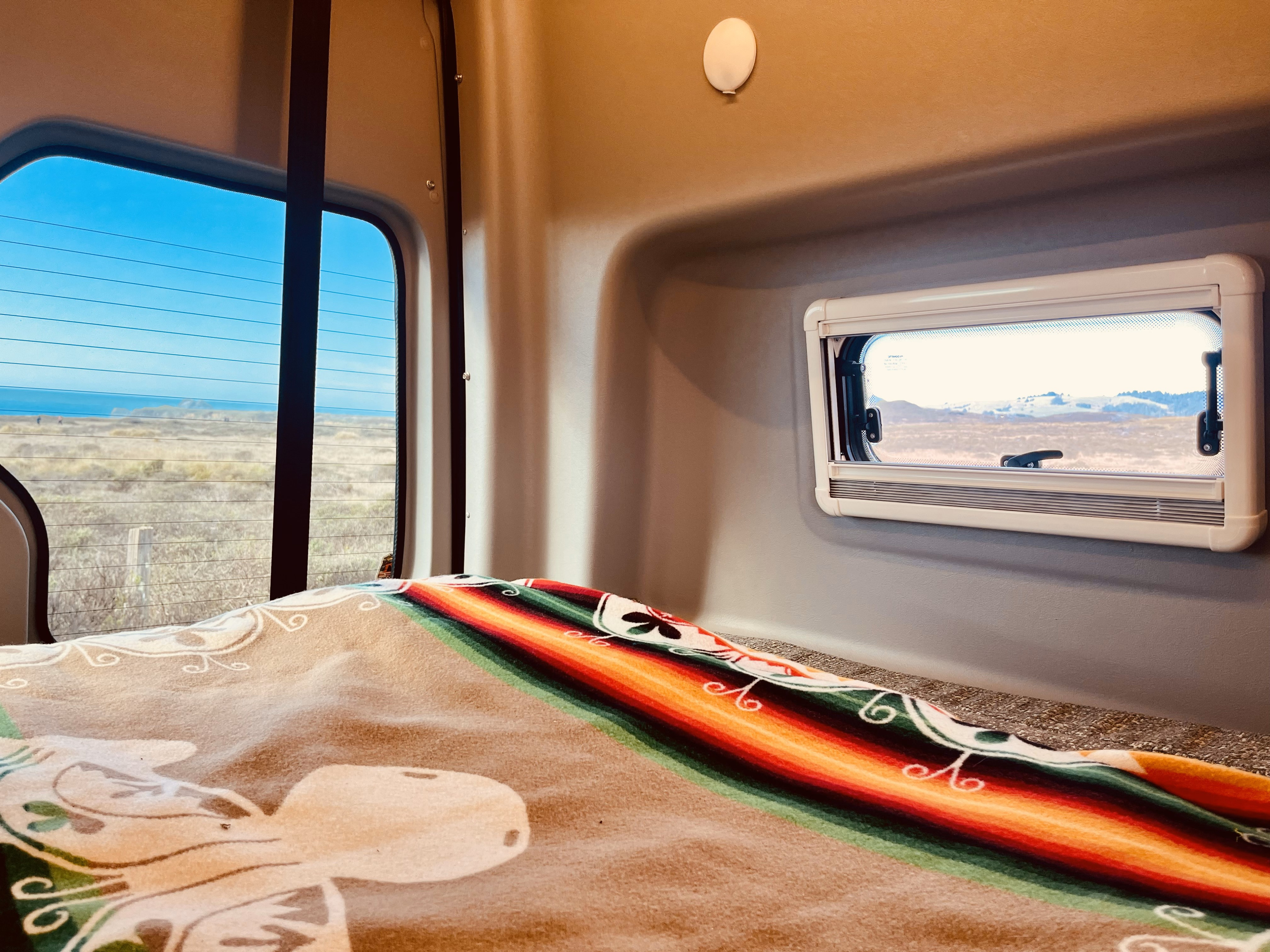 Relax in this RV rental's cozy interior, featuring a colorful blanket and distinct window views of coast and mountains.. Winnebago Revel 2019